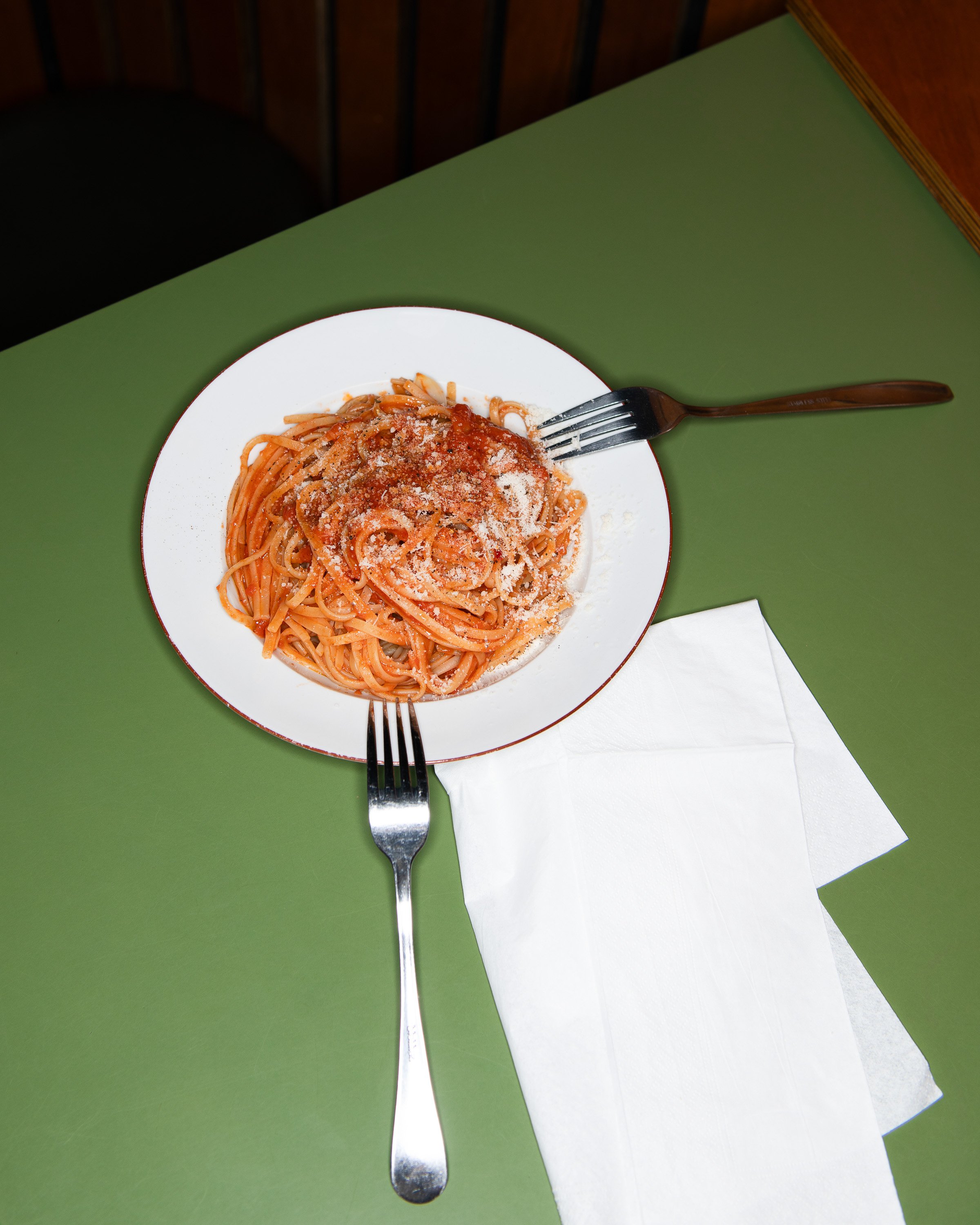 Une assiette de spaghetti à la sauce tomate, garnie de fromage râpé, avec deux fourchettes sur une nappe blanche et un fond vert.