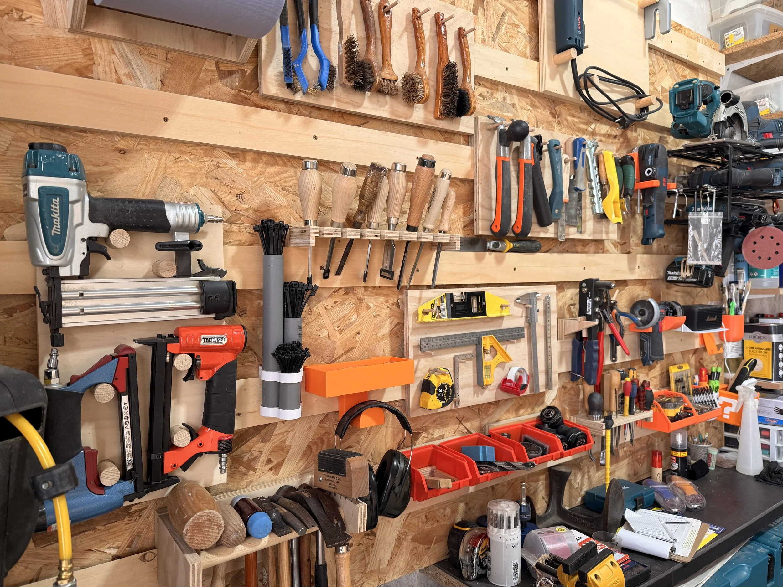 Un mur de rangement en bois avec divers outils de bricolage accrochés, notamment des pinceaux, des tournevis, une scie électrique, une perceuse, des pince coupantes, des niveaux, et des boîtes de rangement avec des accessoires.