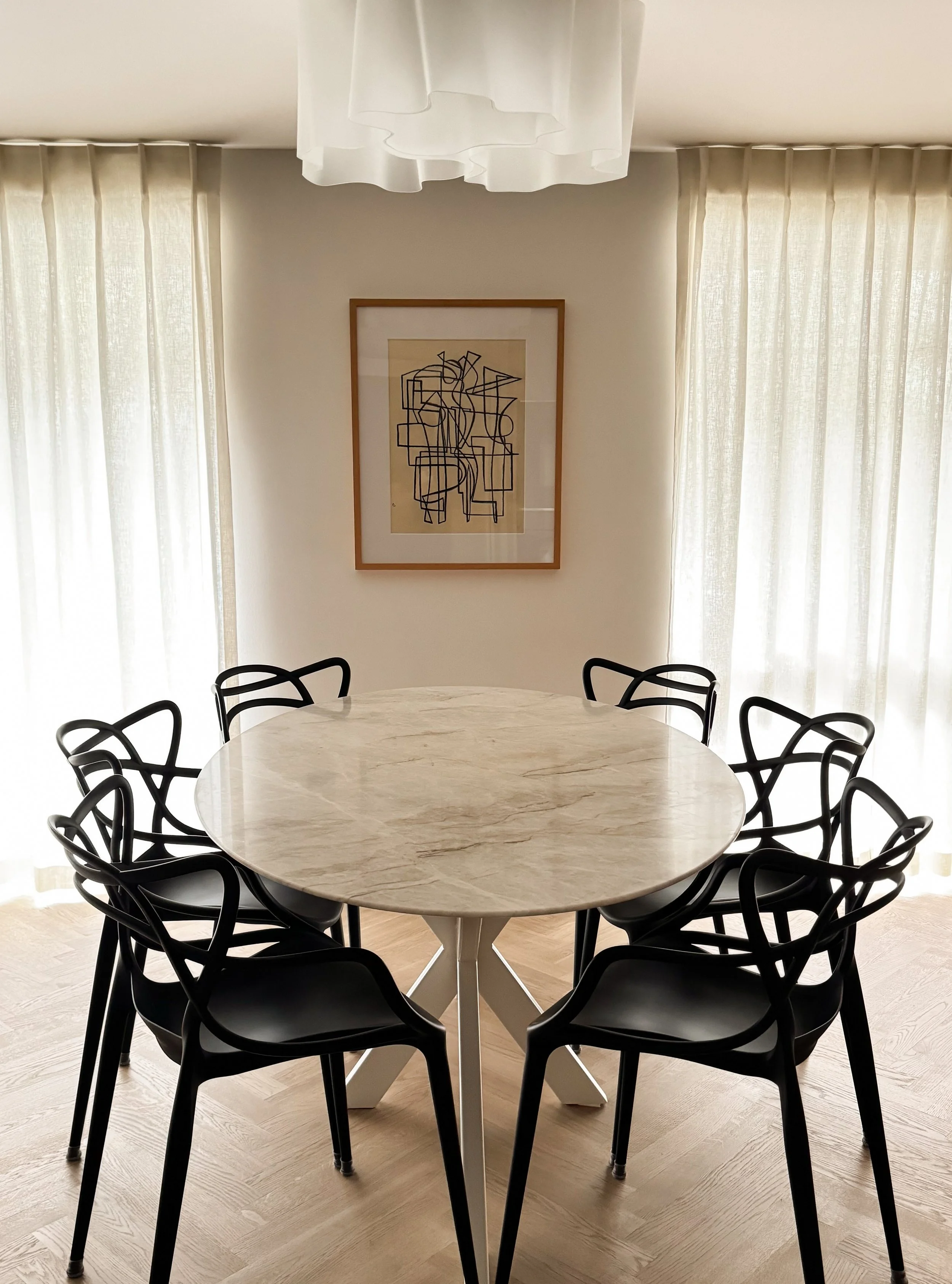 A dining room featuring a round marble table surrounded by black modern chairs with curved backs. The room has cream-colored curtains on two windows, a framed abstract line drawing on the wall, white curtains hanging from the ceiling, and light woode