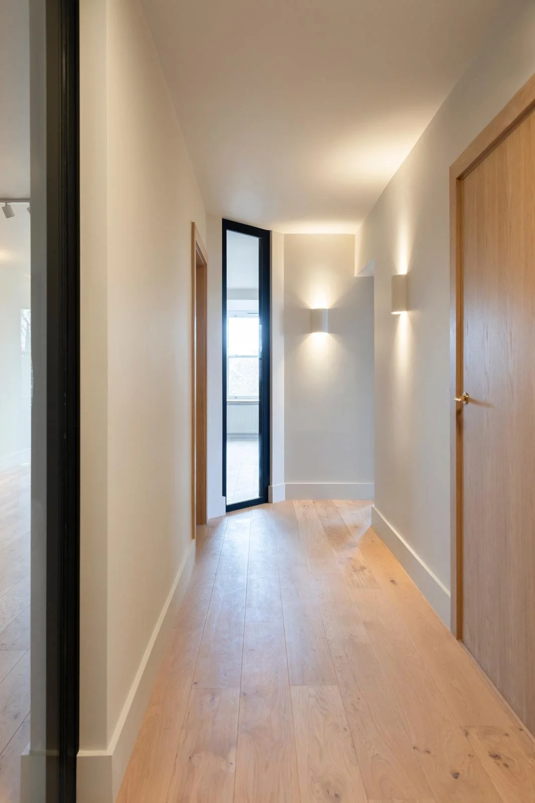 A hallway with wooden floor and white walls, featuring wall-mounted lights and a door at the end leading outside.