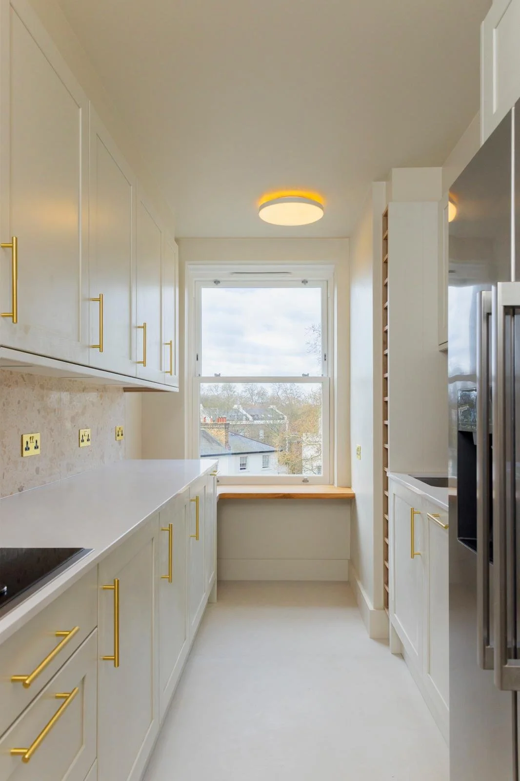 A bright white kitchen with gold hardware, a window at the end, and a built-in wooden wine rack on the right side.