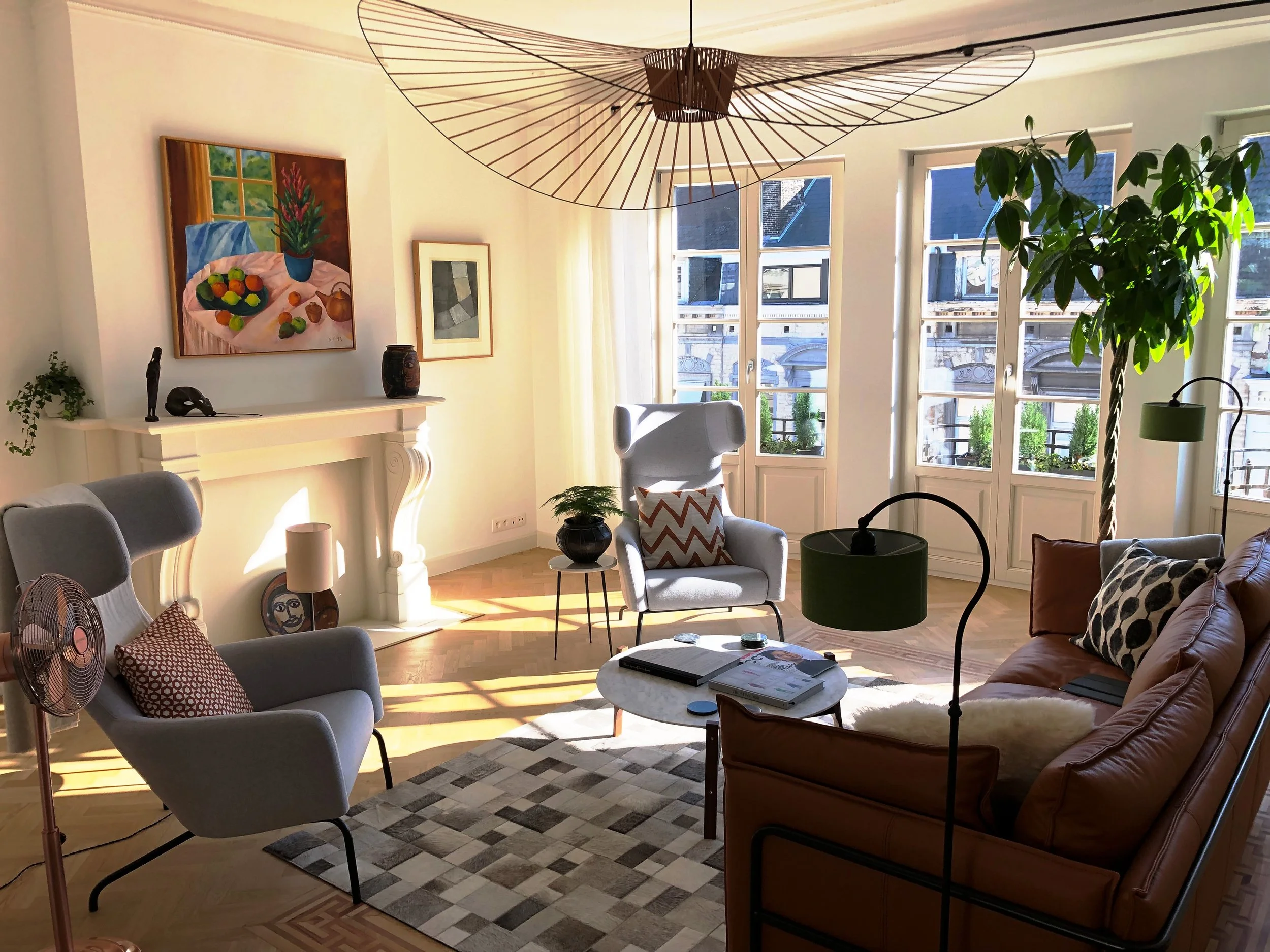 Bright living room filled with natural light from large windows. Contains gray armchair with patterned pillow, tan leather sofa, round coffee table with magazines, and various plants. Decor includes paintings, a lamp, and vases. A contemporary wooden