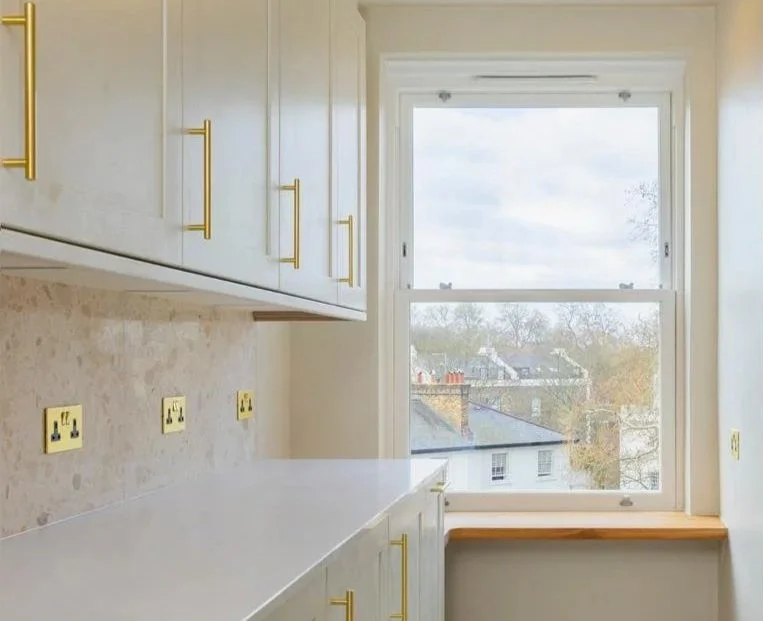 Kitchen with white cabinets, gold handles, a white countertop, and an open window overlooking houses and trees outside.