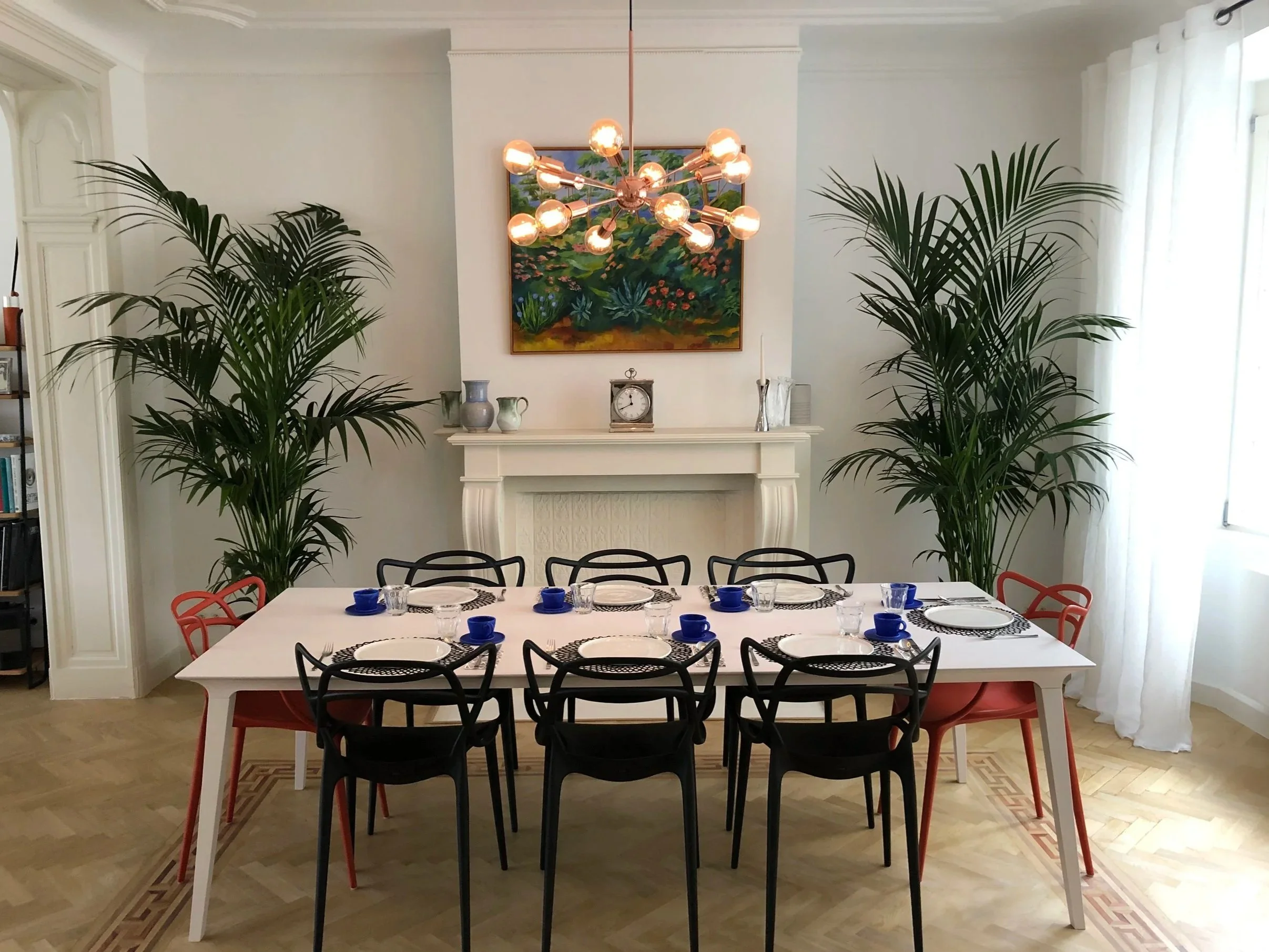 A dining room with a large white table set with black, blue, and white dishware, surrounded by decorative black and red chairs. The room features a white fireplace mantel decorated with vases, a clock, and a candle. Large green plants flank the mantel. A colorful painting hangs above the fireplace, and a modern chandelier with multiple exposed bulbs hangs from the ceiling, centered in the room.