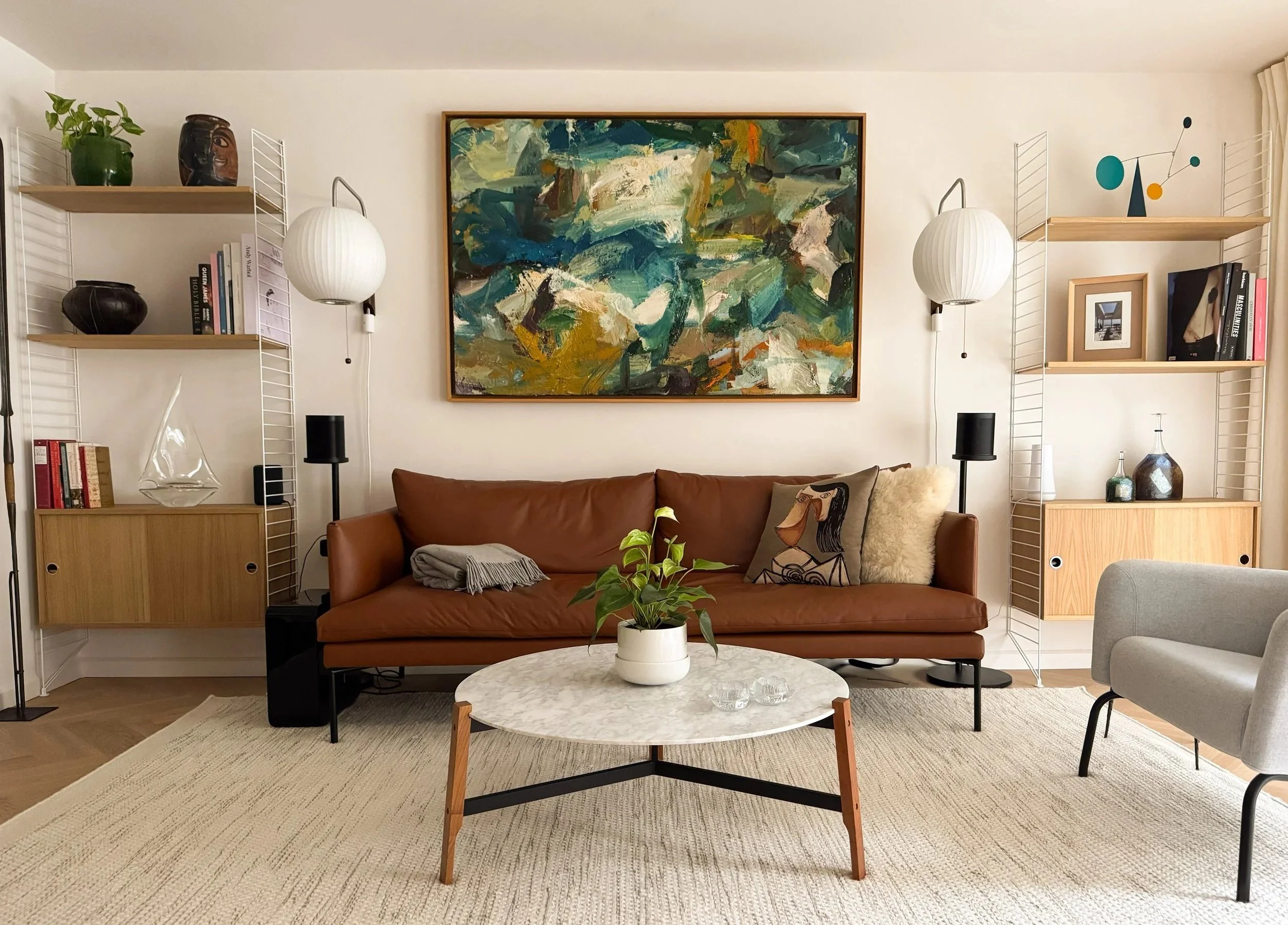 Living room with a brown leather sofa and a round marble coffee table with a potted plant. Abstract painting on the wall behind the sofa, flanked by two white wall-mounted lamps. Shelves with books, vases, and decorative items on either side of the painting.