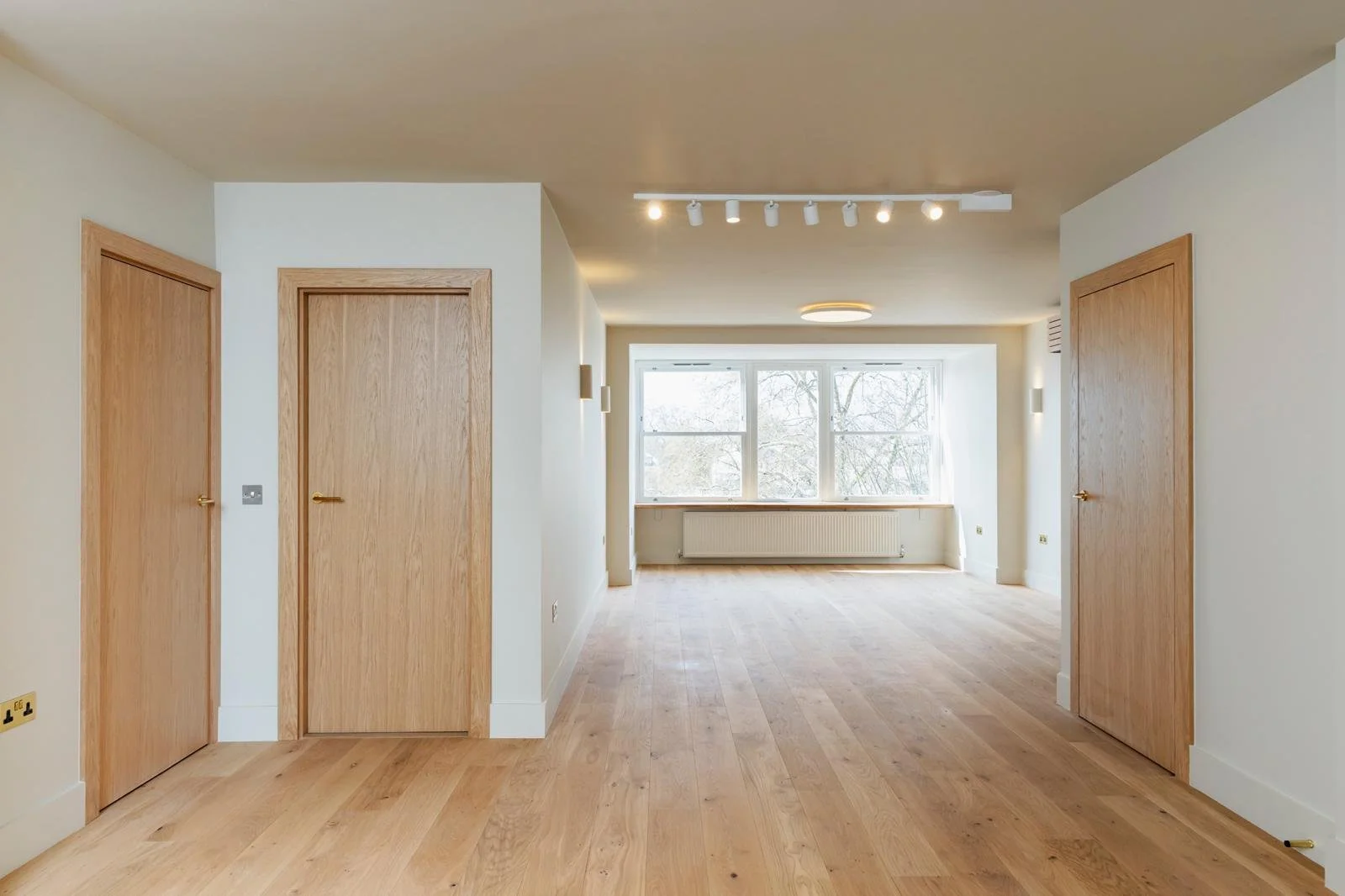 Empty living space with large window, wooden floors, white walls, and two closed wooden doors.
