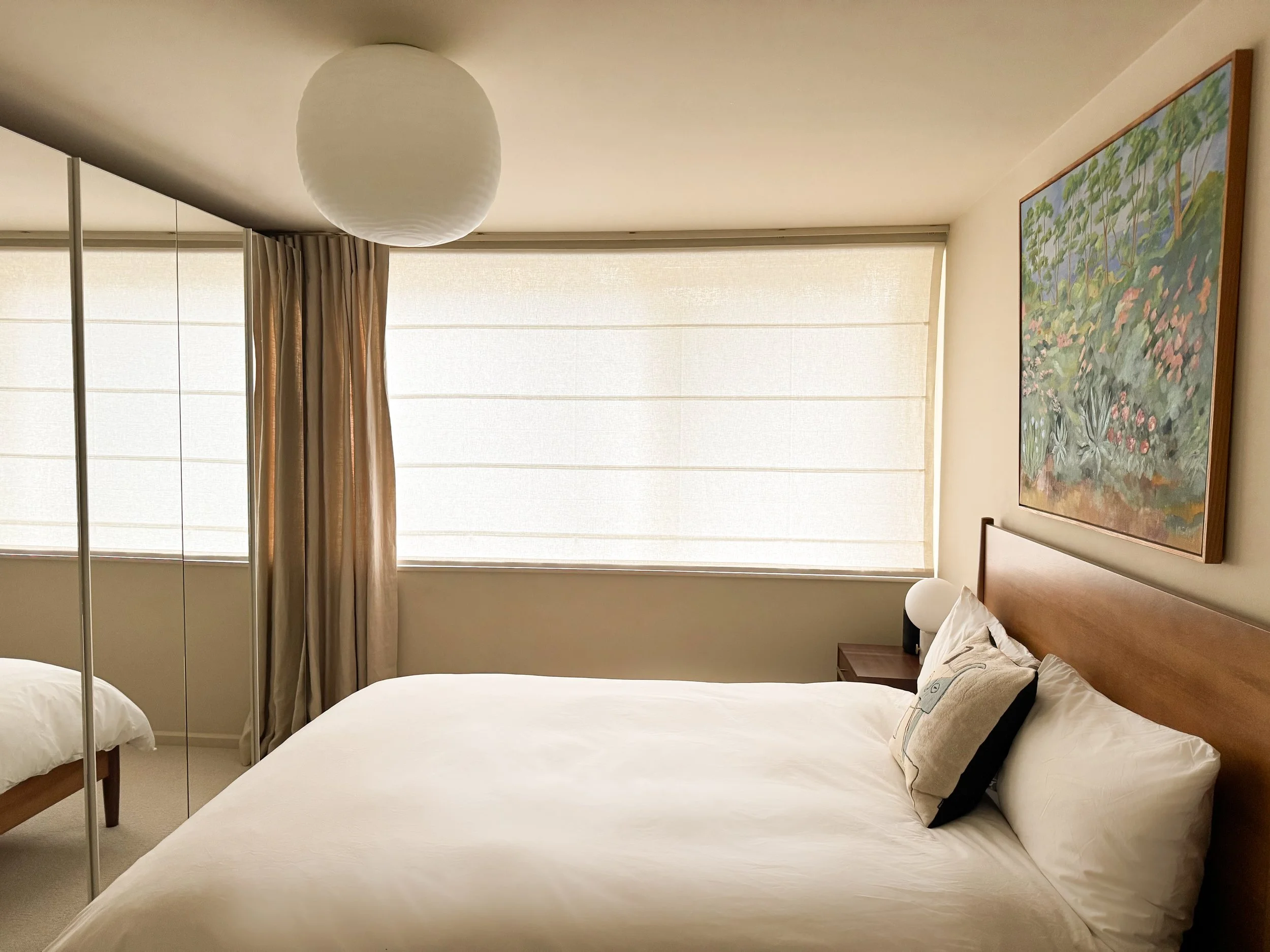 A bedroom with two beds, a nightstand with a lamp, a large painting of a garden scene on the wall, a window with sheer blinds, and beige curtains.