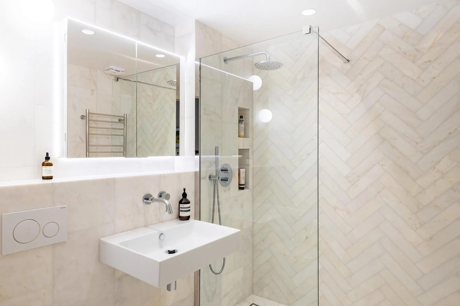 Modern bathroom with a large mirror, white sink, wall-mounted faucet, and walk-in shower with glass partition. The shower area has built-in shelves, a rainfall showerhead, and a textured tiled wall in a chevron pattern.