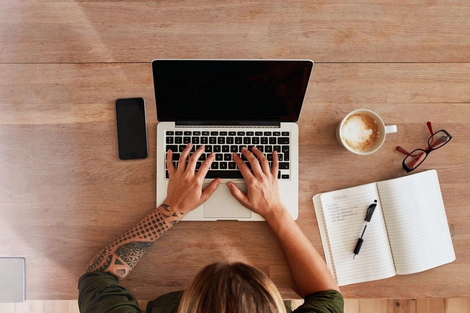 Overhead view of a woman typing on a laptop with coffee, glasses and a to-do list — illustrating the next step women take when they decide to stop smoking for good