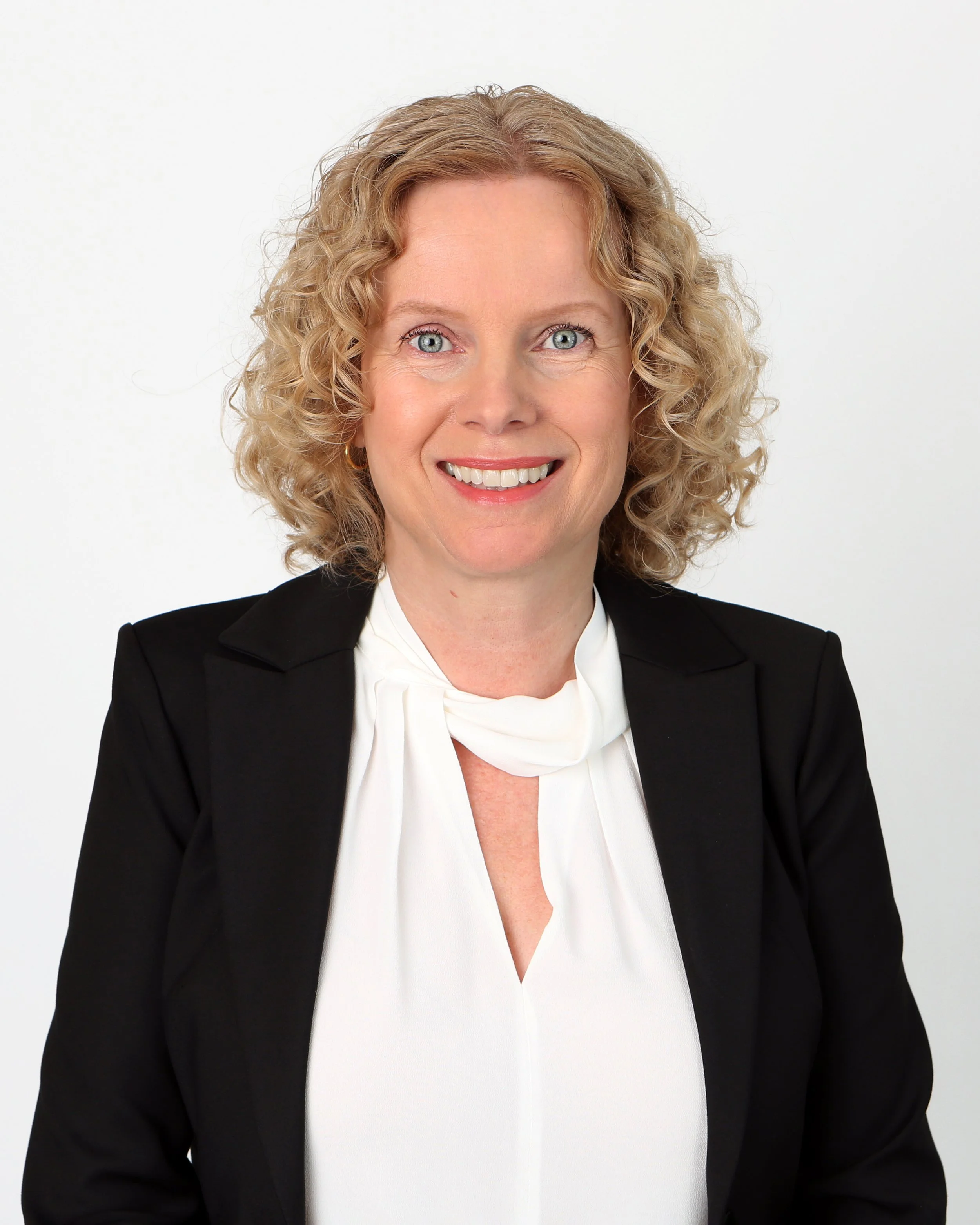 Portret van een vrouw met golvend blond haar, blauwe ogen, glimlachend, gekleed in een zwart colbert en een witte blouse, tegen een witte achtergrond.