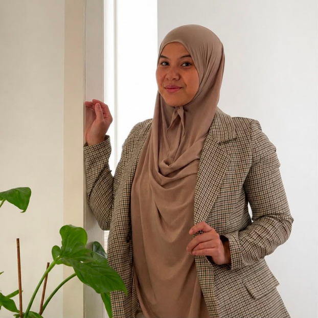 Vrouw met hoofddoek en nette blazer staat naast een plant in een kamer met witte muren.