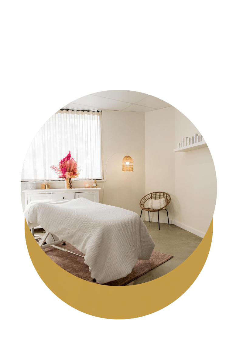 Une pièce de massage ou de relaxation avec un lit recouvert d'une couverture blanche, un fauteuil en rotin avec un coussin, une lampe suspendue et un bouquet de fleurs séchées sur une console blanche.