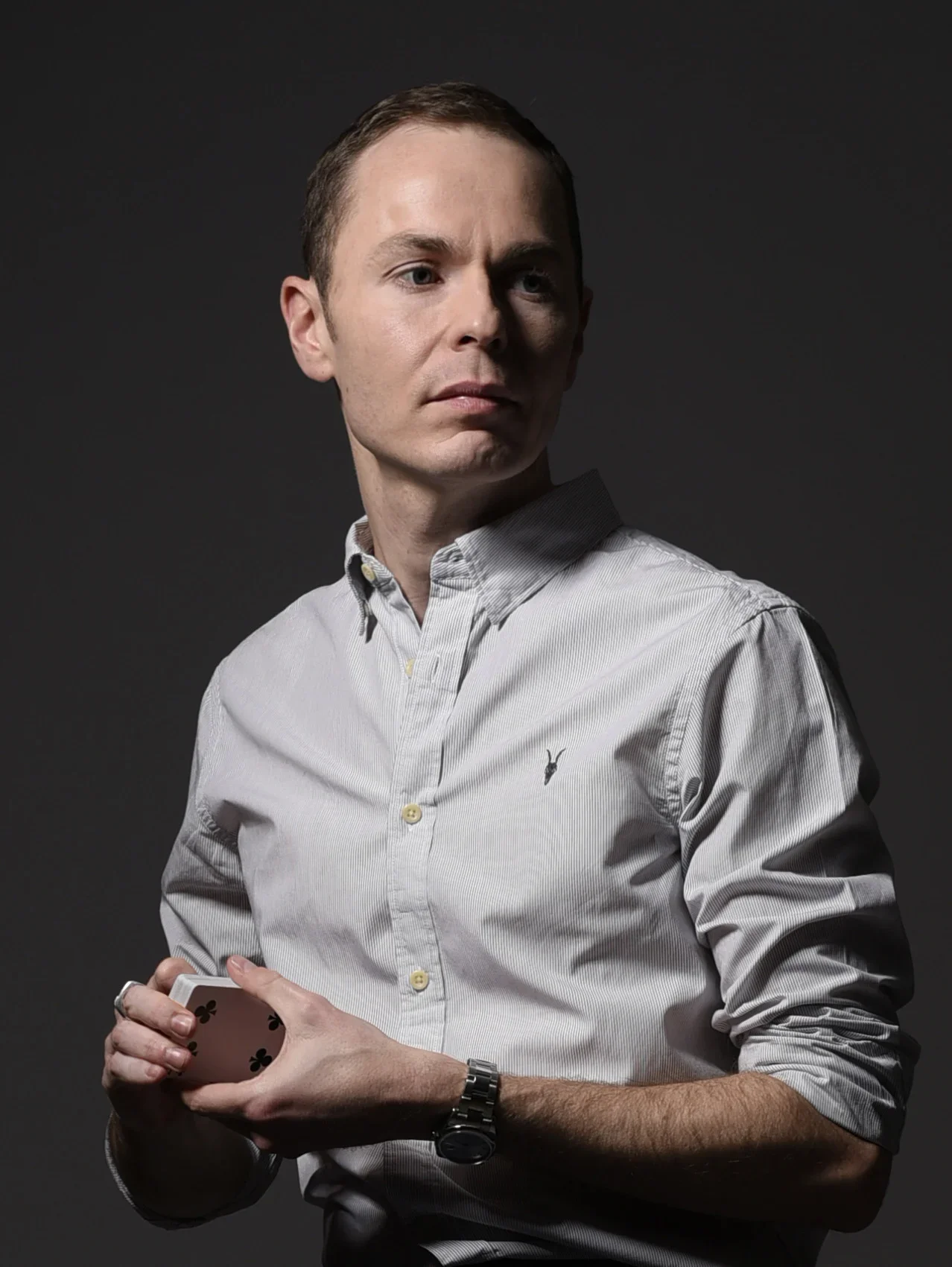 Magician, Martyn Rowland wearing a light grey button-up shirt with a small embroidered logo, holding playing cards in his right hand, standing against a dark background.