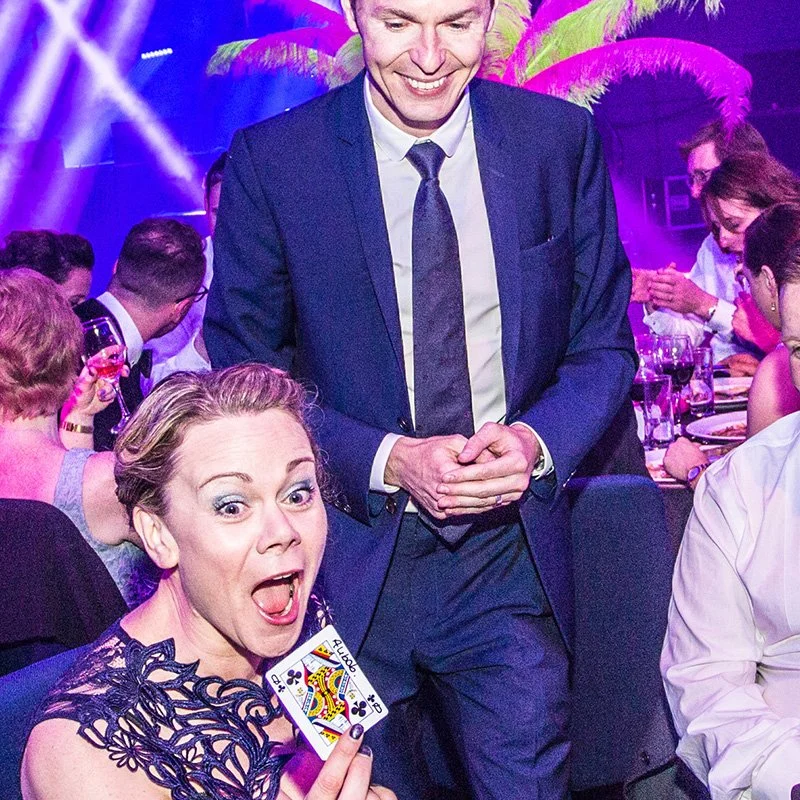 A woman at a festive event showing a playing card, the Queen of Clubs, with an excited expression, while Martyn, in a suit stands beside her, surrounded by other people at a table with drinks under colourful lighting.