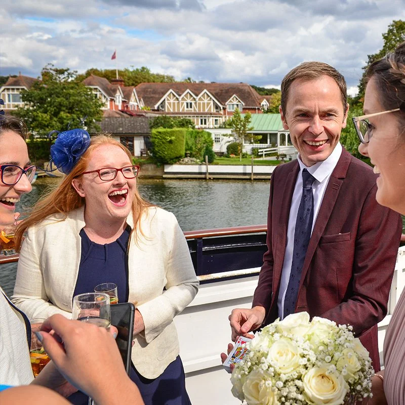 People enjoying a boat ride on a river, smiling and laughing, with a view of houses and trees along the shoreline under a partly cloudy sky.