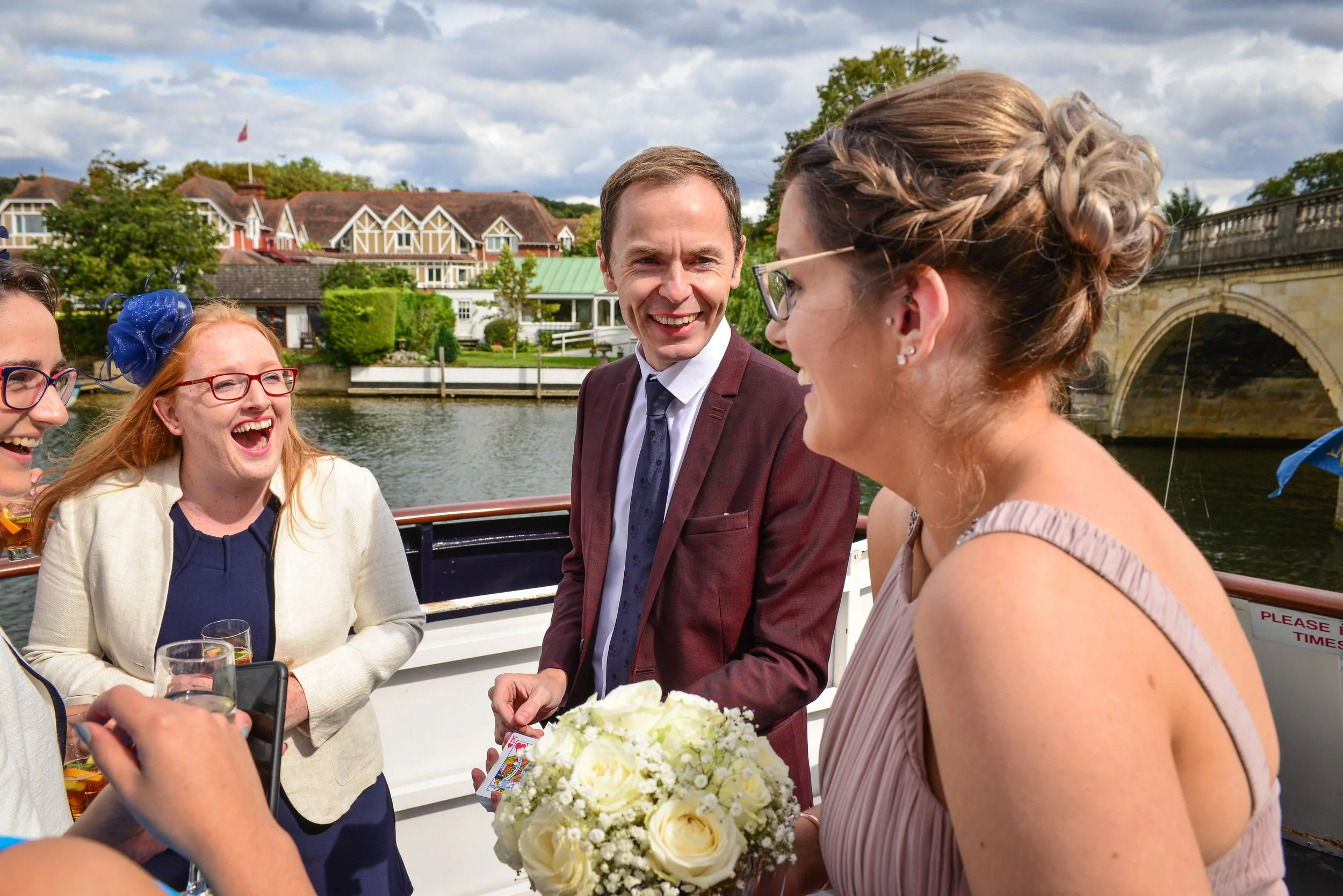 People smiling and celebrating on a boat, with a woman holding a bouquet of white roses, and Martyn in a suit. Buildings and a bridge are in the background under a partly cloudy sky.