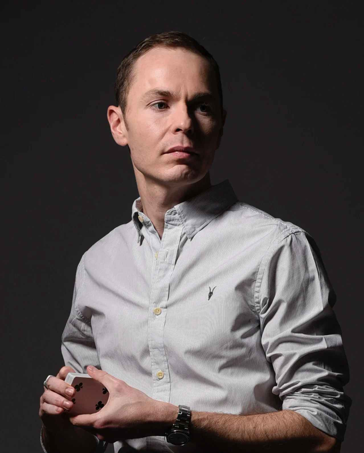 Magician, Martyn Rowland, wearing a light-coloured button-up shirt, holds a deck of playing cards in his left hand, standing against a dark background.