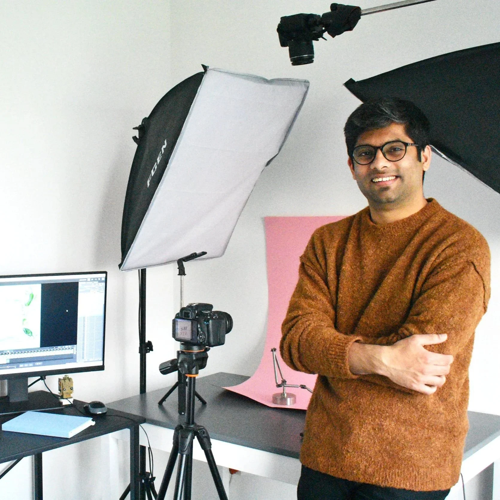 A man with glasses, wearing a brown sweater, standing in a photography studio with equipment, softbox lights, a camera on a tripod, a computer monitor, and a pink backdrop.