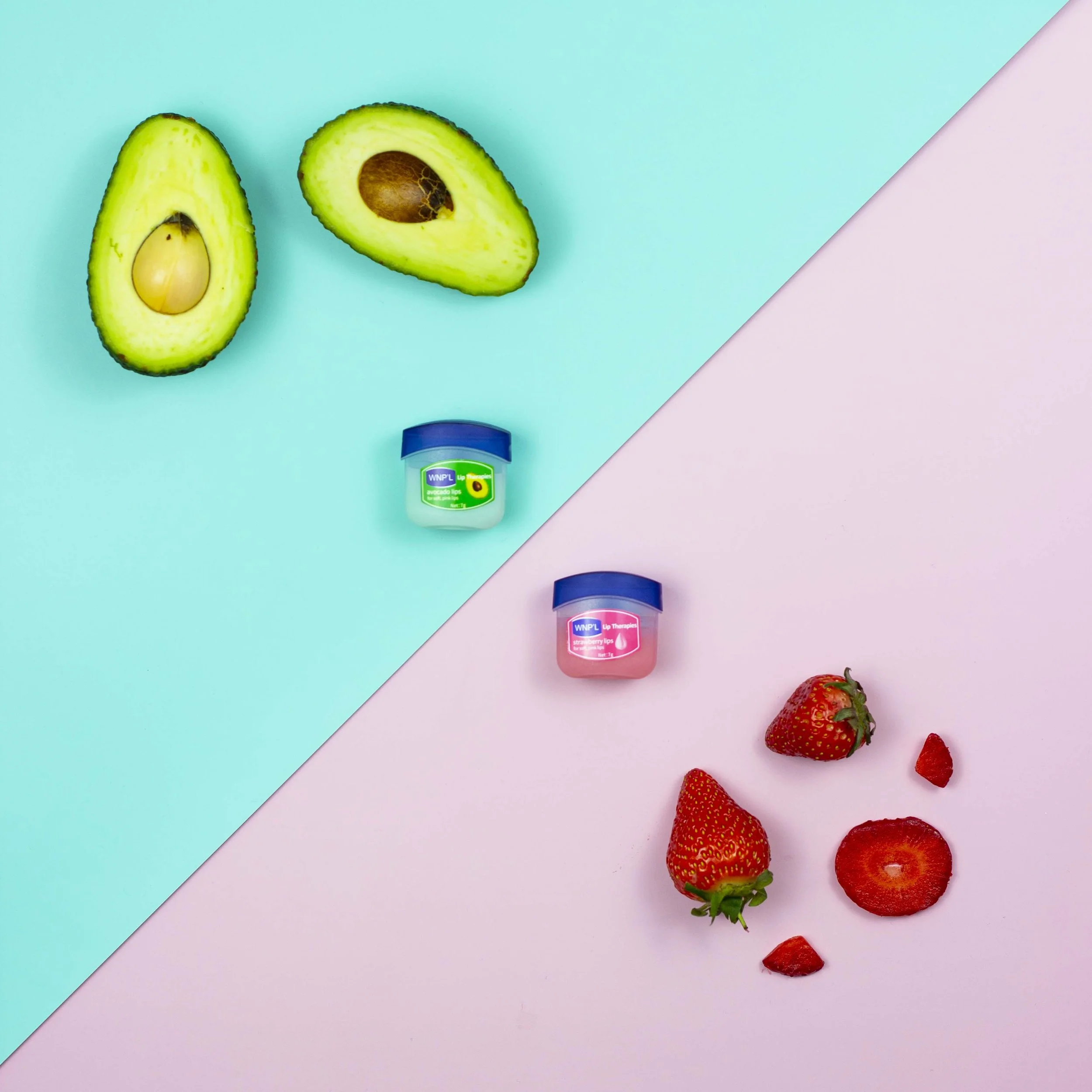 Two halved avocados with one showing the seed and the other with the seed removed, placed on a split pastel blue and pink background. Below the avocados, there are two small containers of lip balm, one labeled "avocado" and the other "strawberry". To the right, there are fresh strawberries, some whole and some sliced.