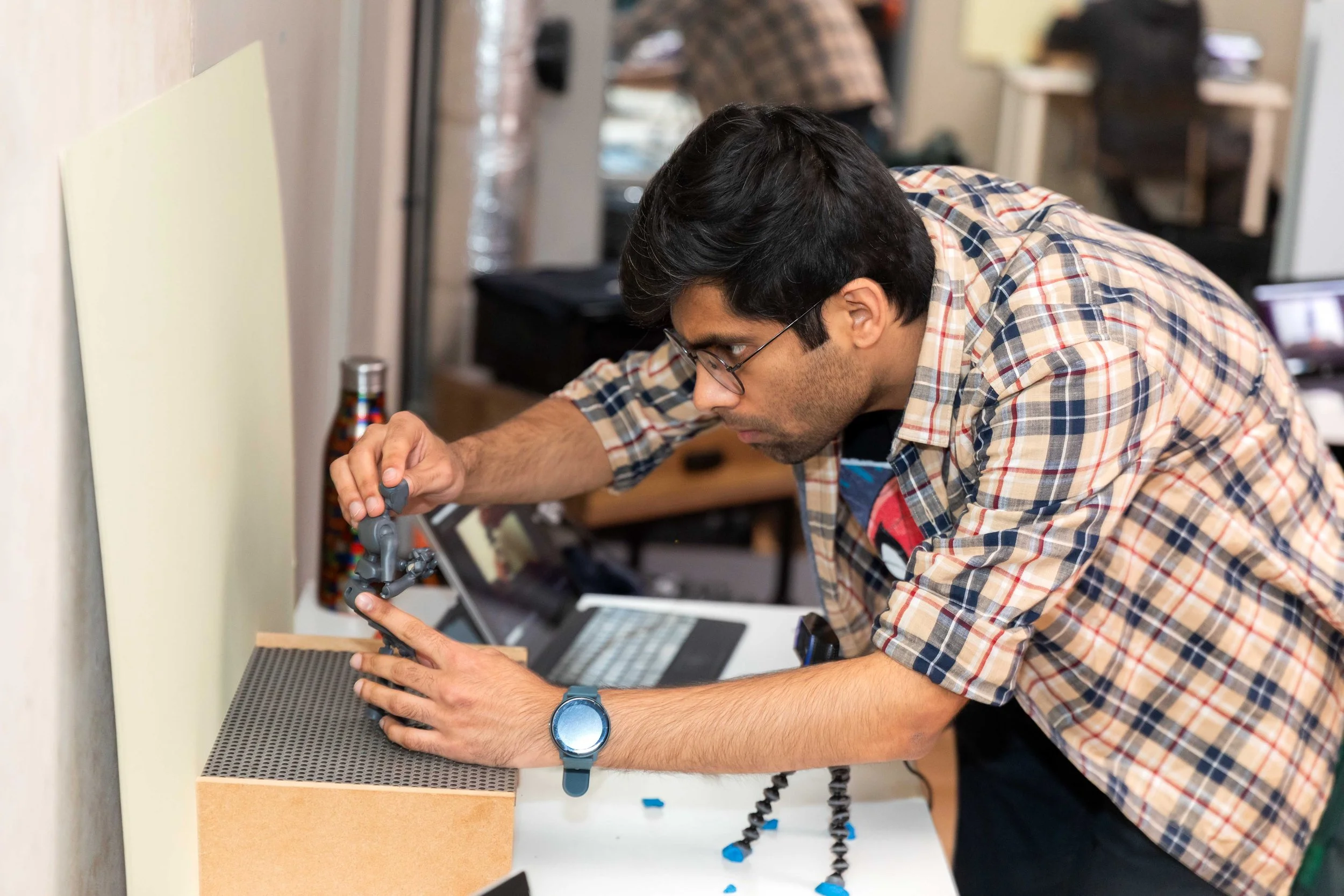 A man wearing glasses and a plaid shirt is engaged in a creative project, working with a flexible arm tool on a small object. He is seated at a workbench in a workshop or lab, with various tools and equipment scattered around him.