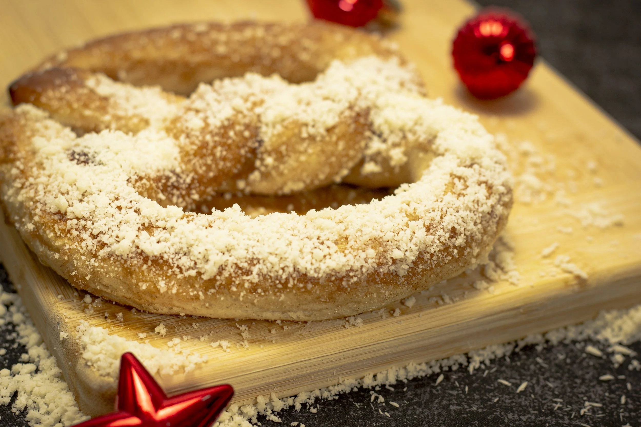 Close-up of a donut with crumbly topping on a wooden cutting board, with a red star-shaped ornament in the foreground and a red object in the background.