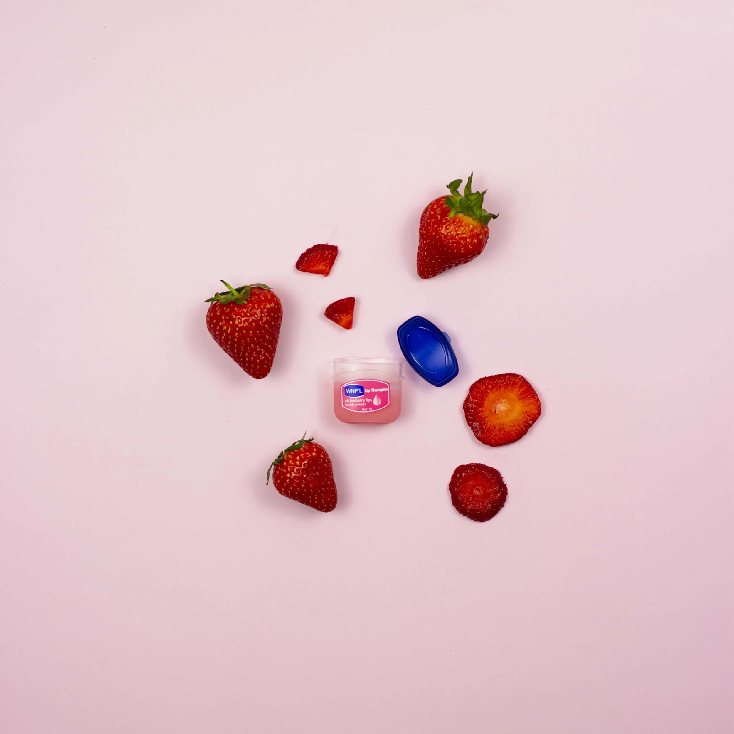 Several strawberries, some halved, scattered on a pink background, with a small bottle of strawberry lip balm and a blue capsule nearby.