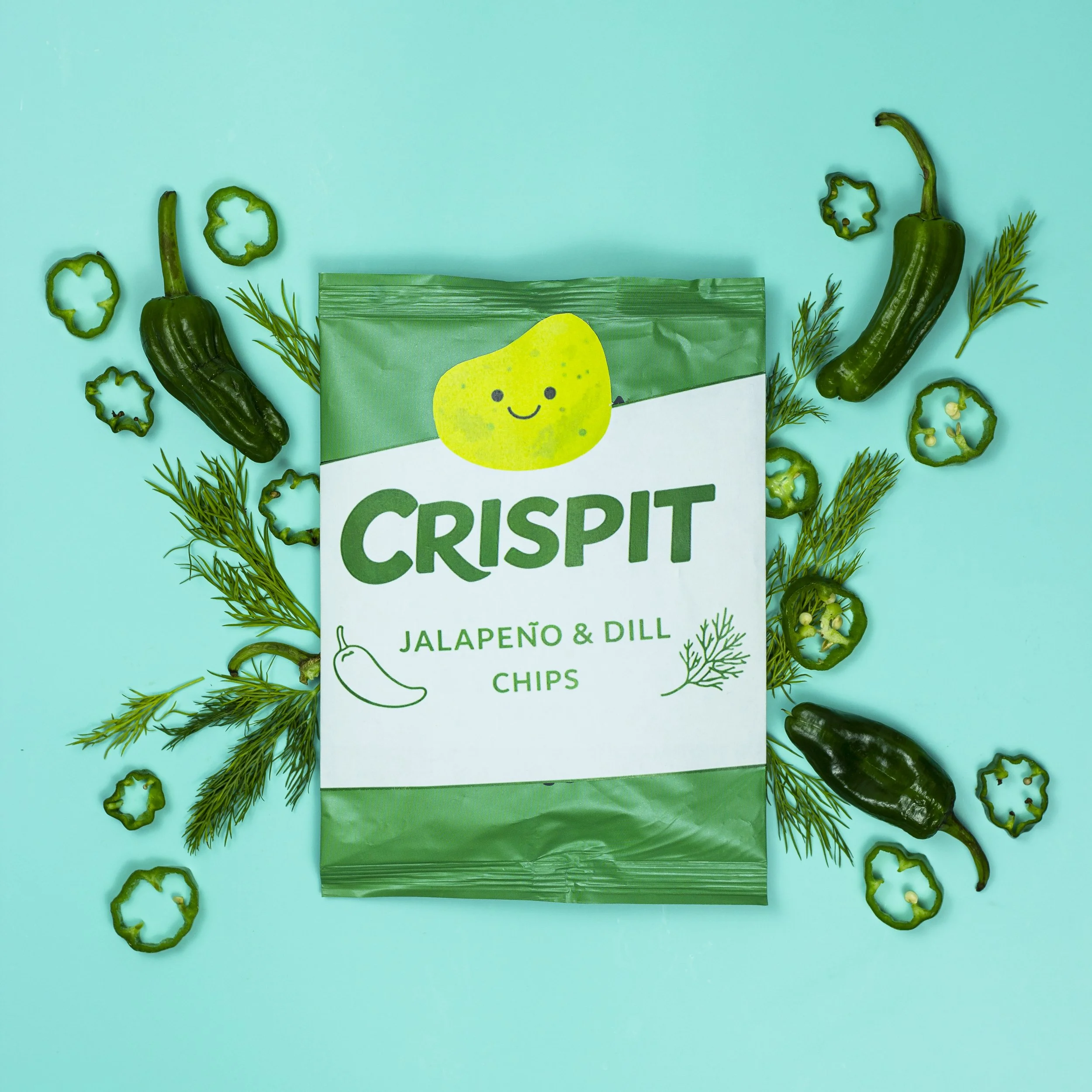 Crispit jalapeño and dill chips packaging with sliced jalapeño peppers and dill sprigs arranged on a teal background.