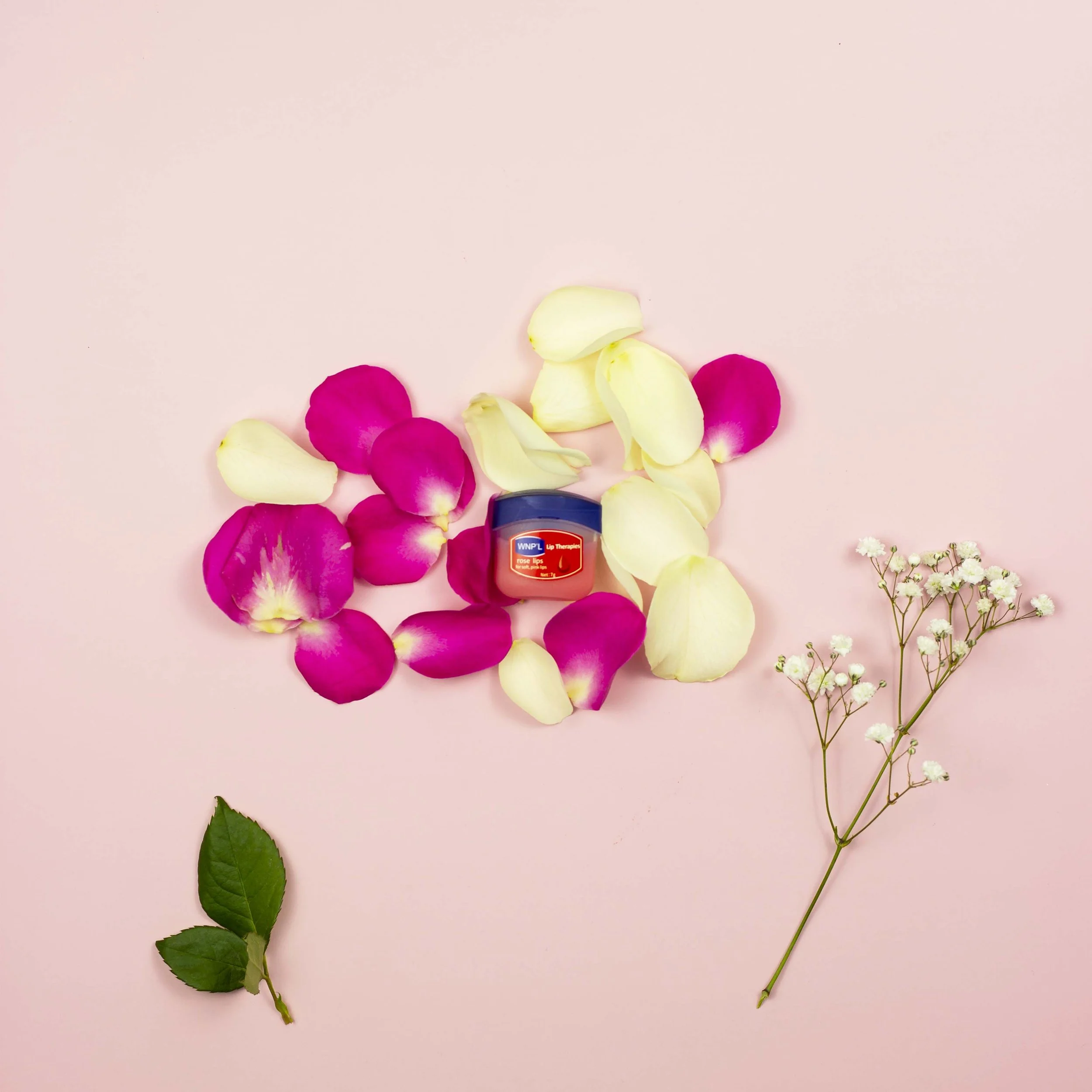 Various flower petals in shades of pink and cream, green leaves, and a bottle of lip balm on a pink background.