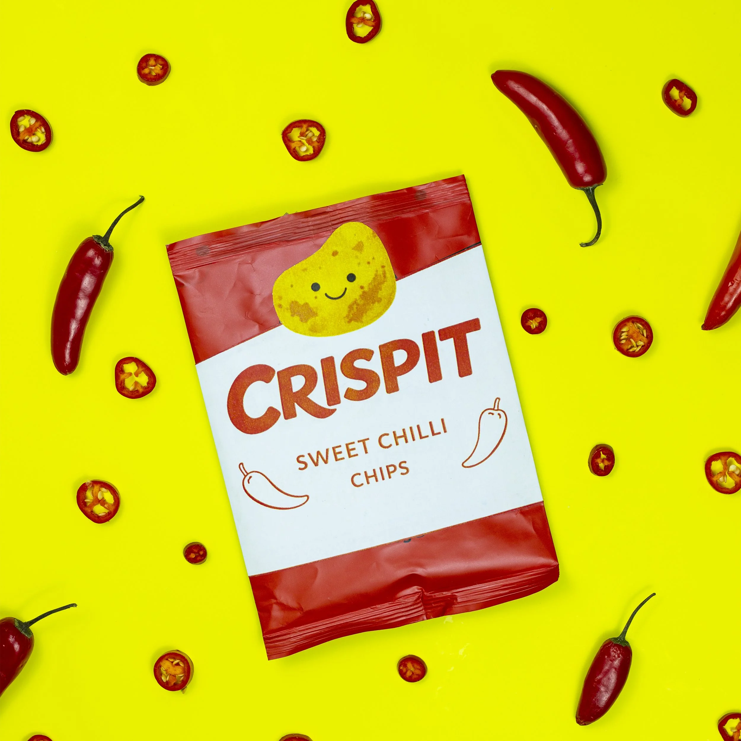 Red chili peppers and slices of red chili scattered on a bright yellow background. There is also a partially visible red and white snack package with a yellow chick illustration and the word "CRISPIT" on it.