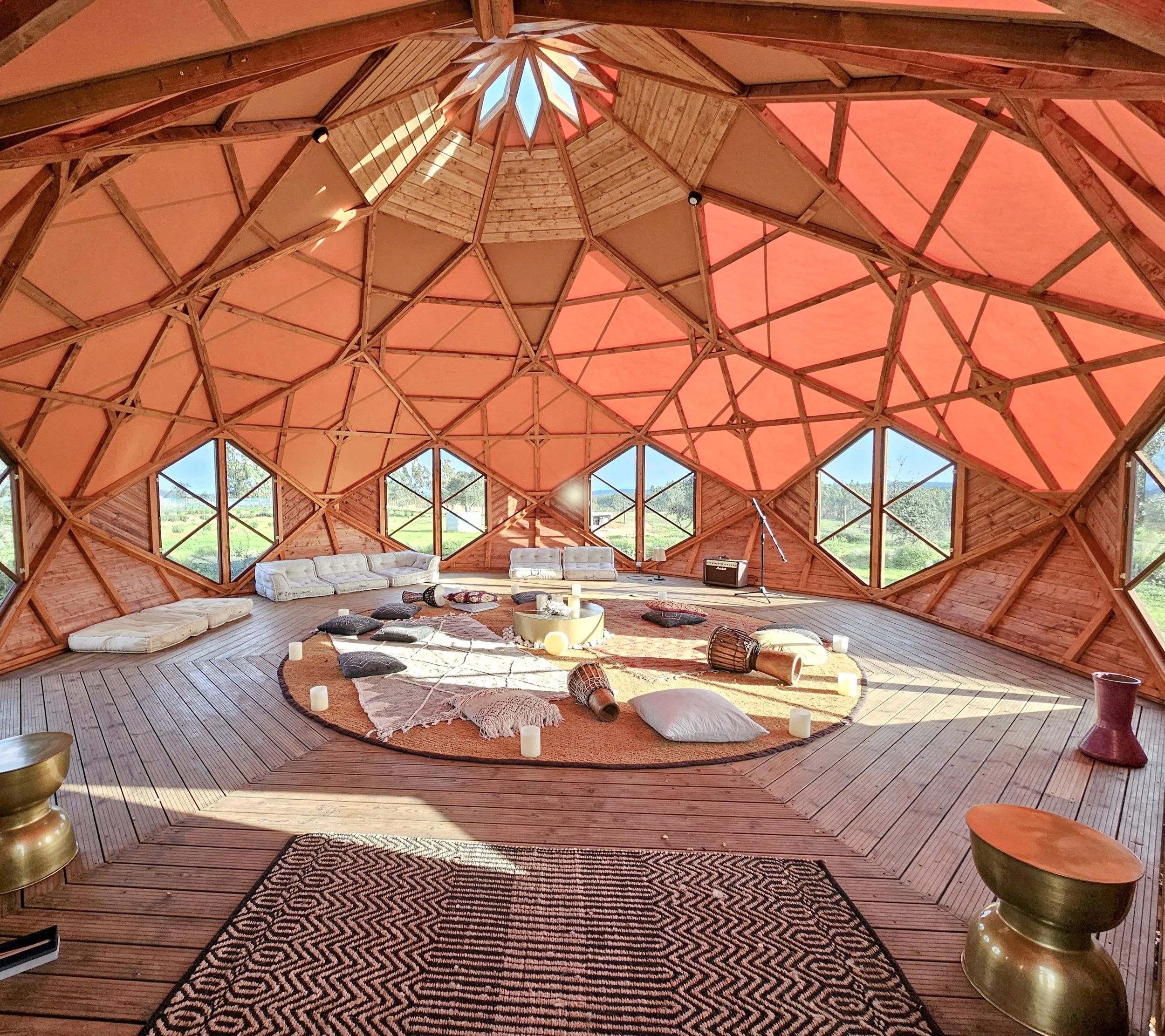Interior view of a geodesic dome with wooden framework, pink fabric ceiling, and large windows showing a green landscape. The floor is wooden, with cushions and a round area rug, and various decorative items, including candles and drums, are arranged in the center, creating a cozy meditation or relaxation space.