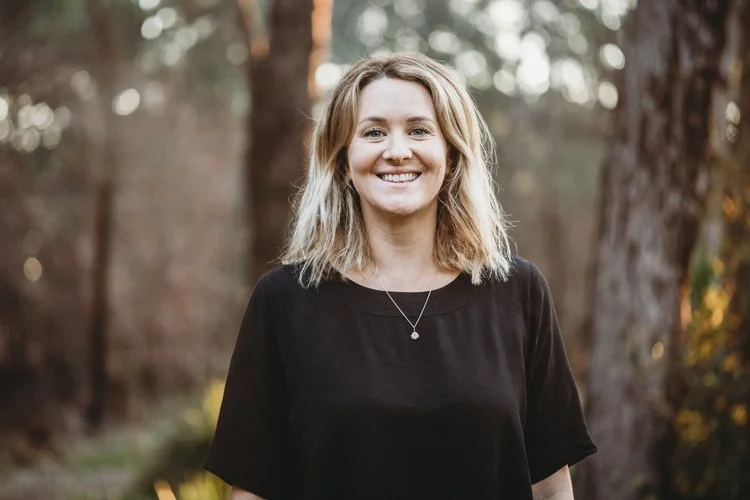A woman with shoulder-length blonde hair smiling outdoors in a wooded area, wearing a black t-shirt and a necklace.
