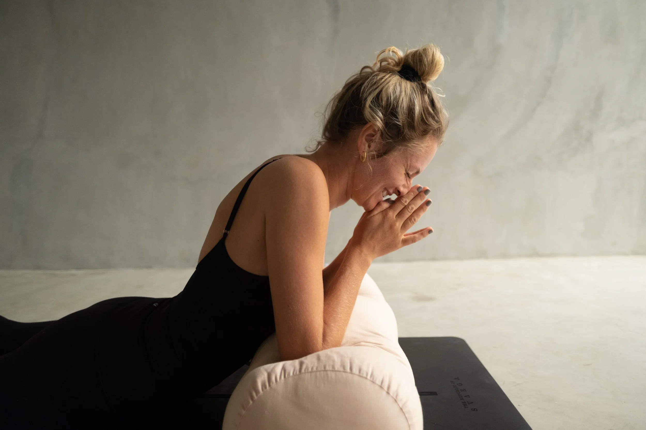 A woman with blonde curly hair tied up in a bun, wearing a black tank top, practicing yoga or meditation with her hands in a prayer position, smiling with eyes closed, in a minimalist room.