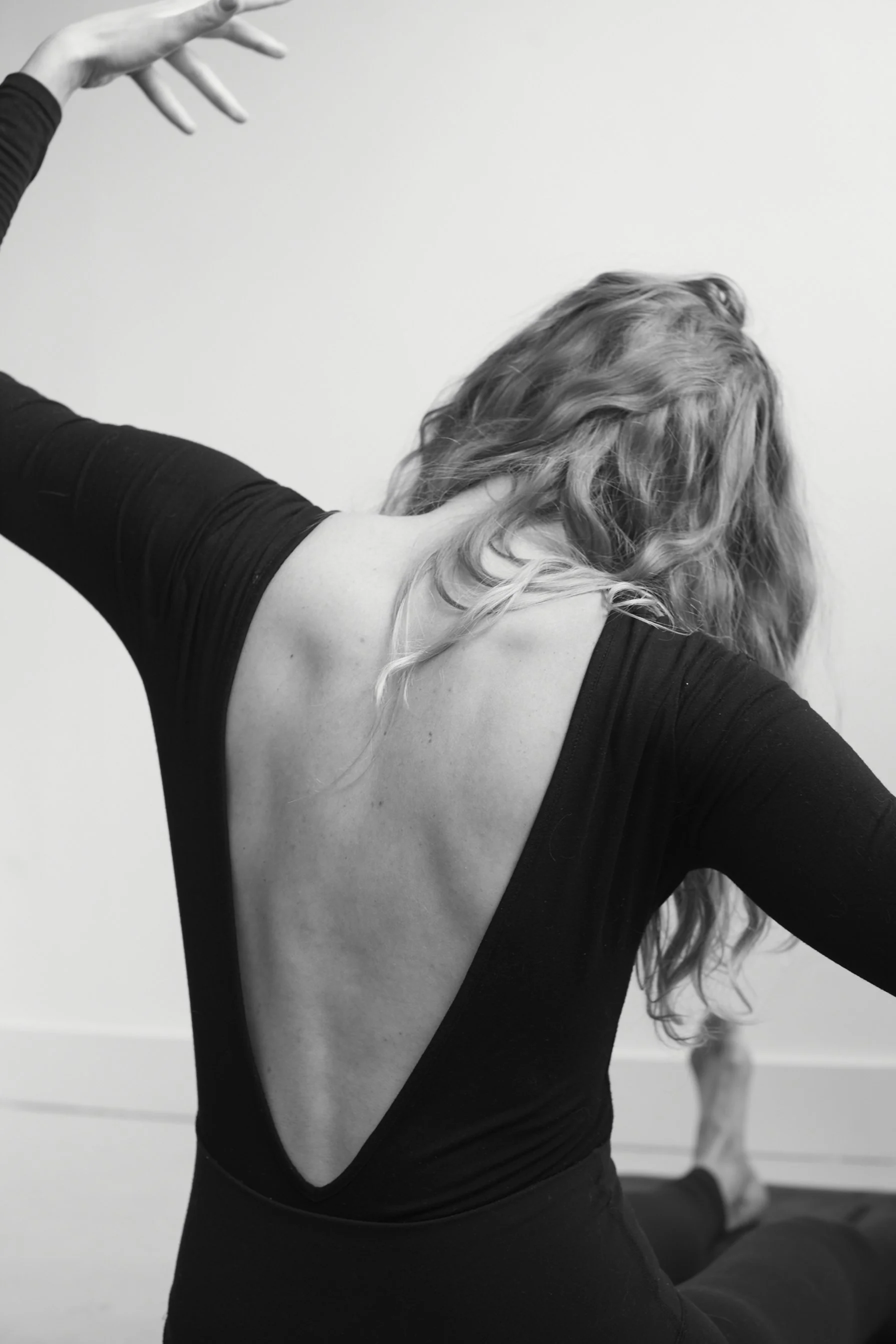 A woman with curly hair practicing yoga, sitting in a pose with one arm extended and the other hand on her neck, wearing a black open-back top, in front of a plain wall.