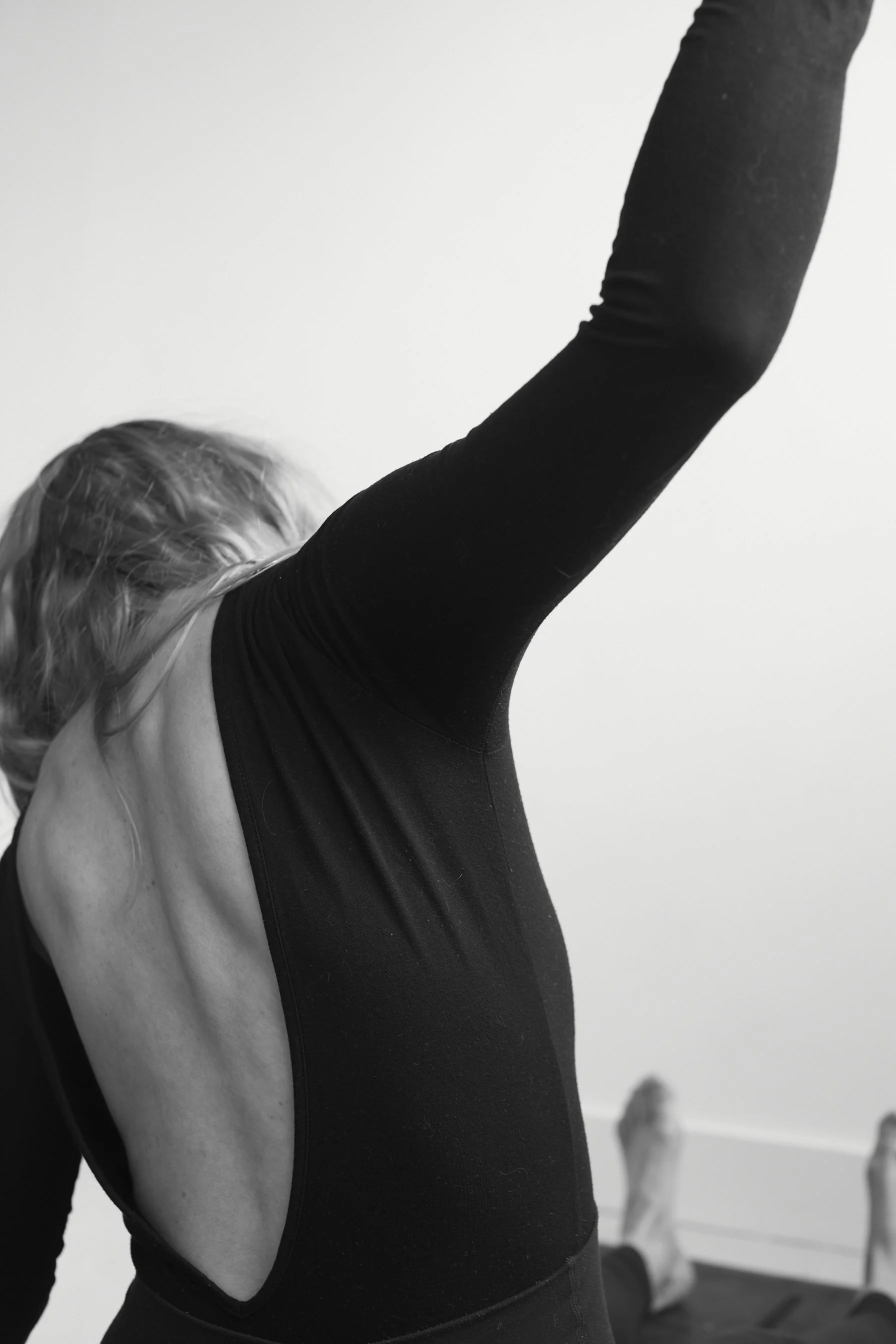 A person with blonde hair wearing a black long-sleeved top is stretching one arm overhead in a room with a plain wall and a piece of furniture in the background.