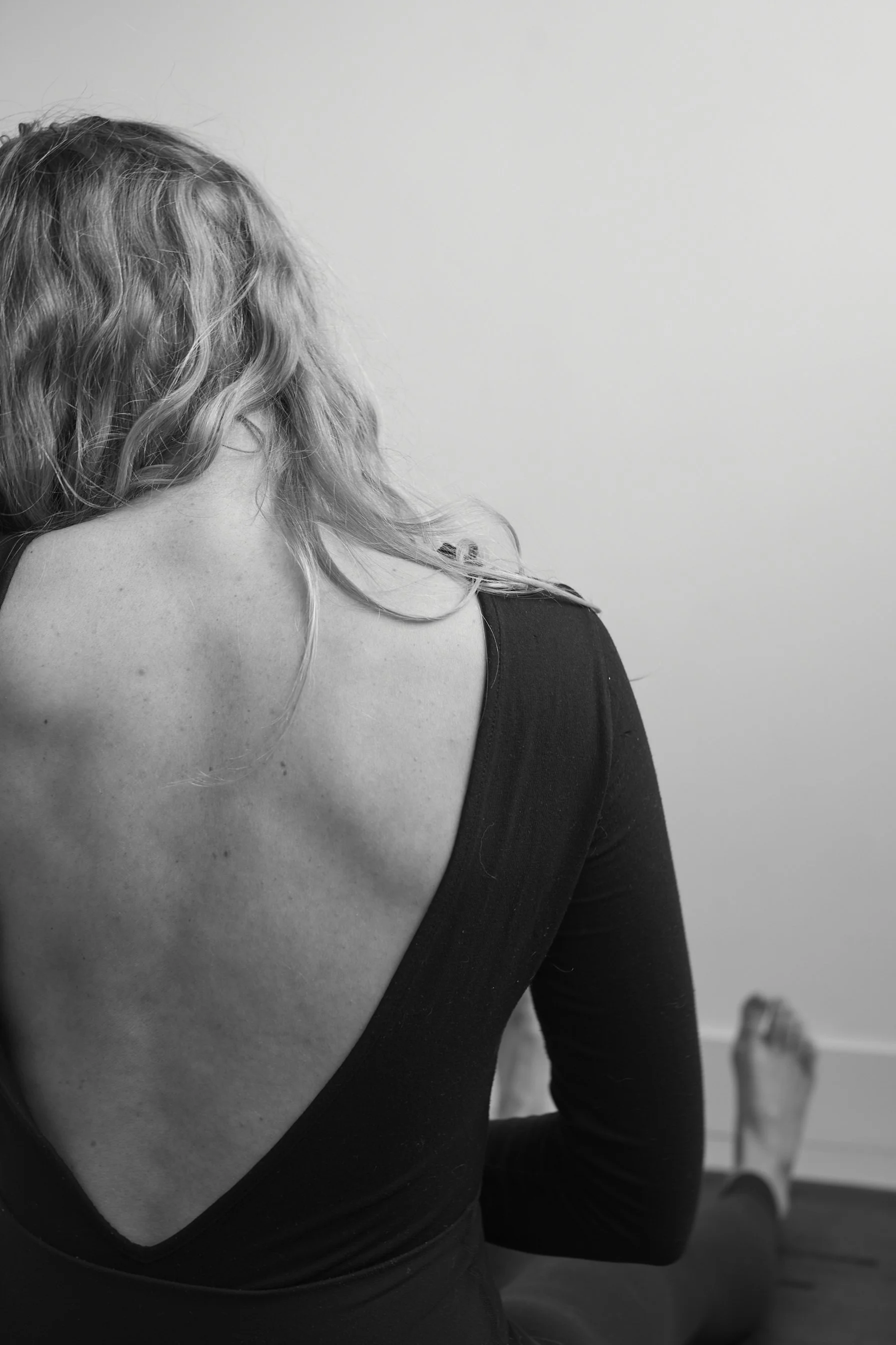 Black and white photograph of a woman with curly hair, wearing a low-cut top, with her back to the camera. She is standing in a room with a plain wall and a chair in the background.