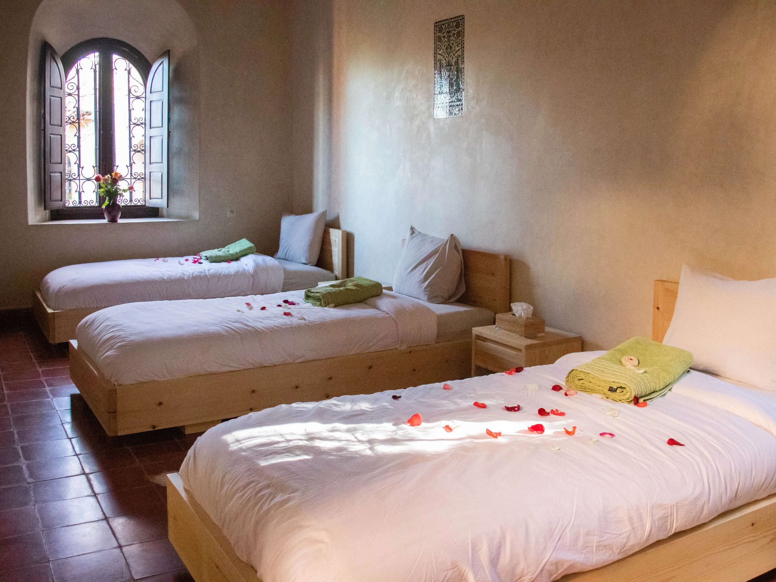 A bedroom with three single beds, each with white sheets, pillows, and towels, decorated with rose petals and flowers. An arched window with open shutters, a vase with flowers on the window sill, and a small wooden nightstand between the beds.