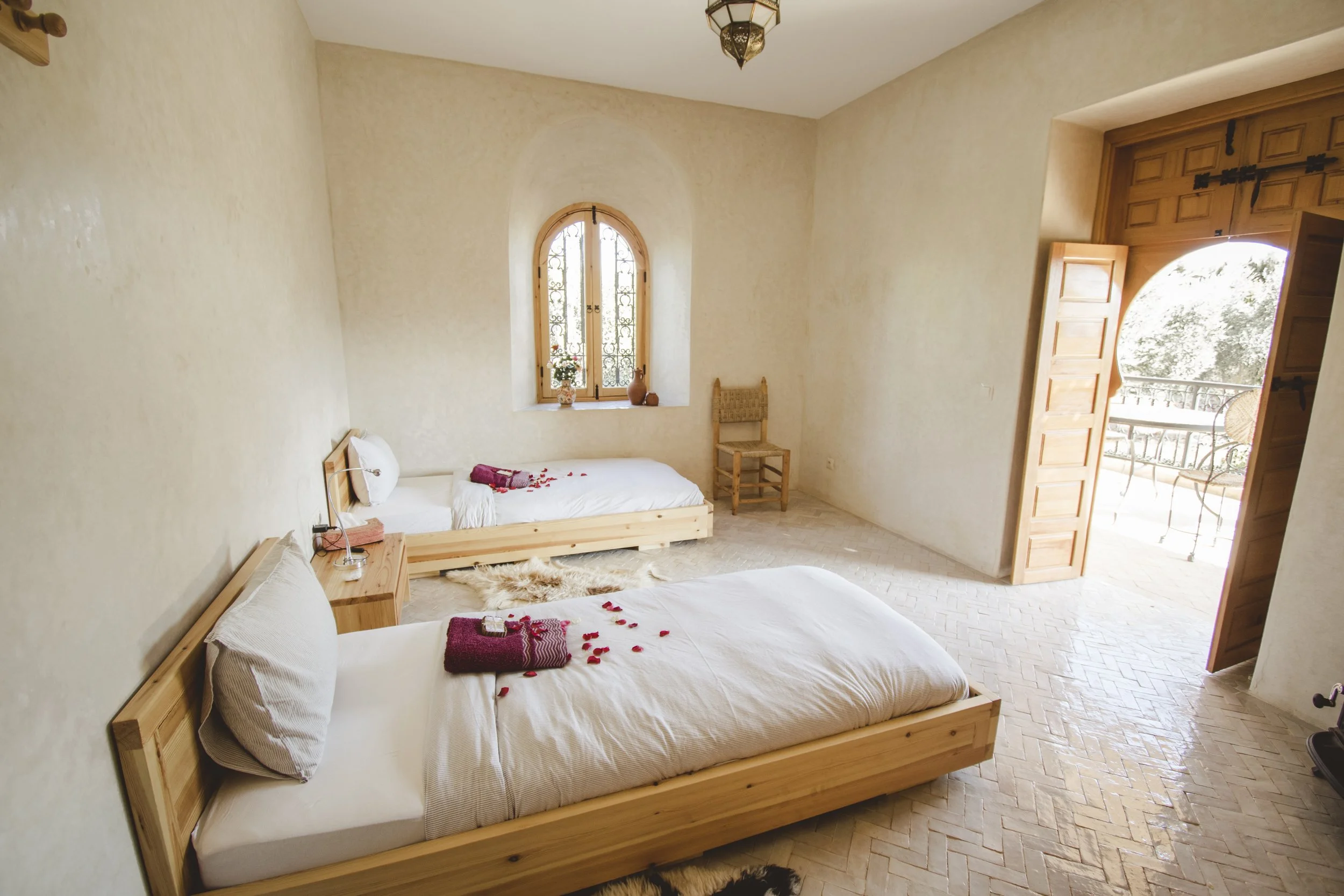 A spacious bedroom with two wooden beds, decorated with towels and rose petals. There is a small window with iron bars, a wooden chair, and an open balcony door with a curved top leading outside. The room has cream-colored textured walls and herringbone-patterned wooden flooring.