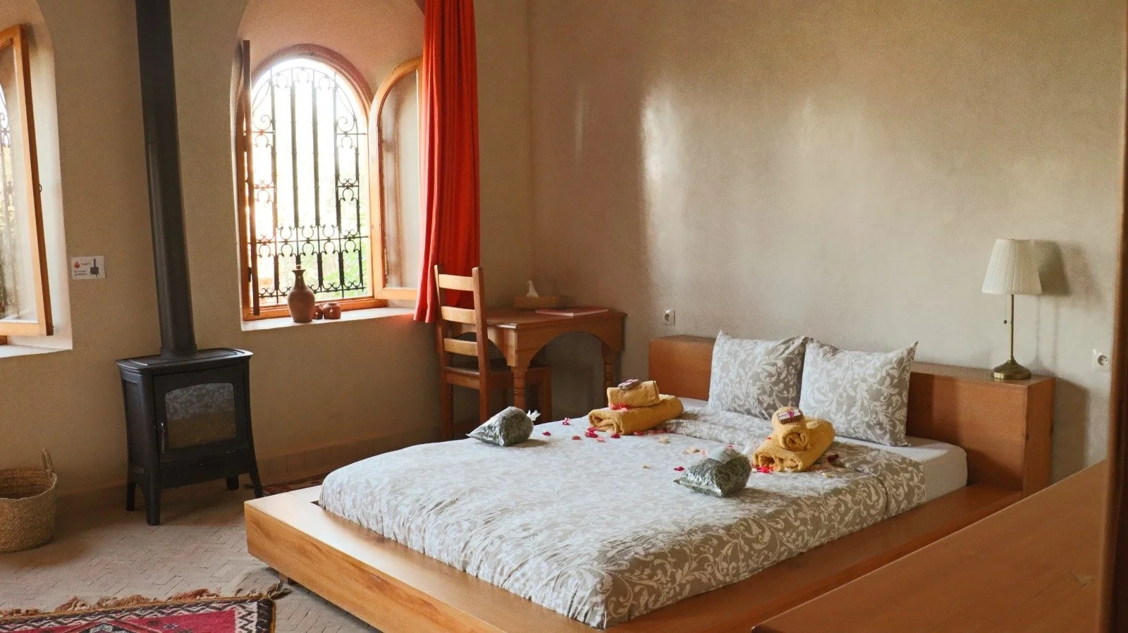A cozy bedroom with a bed adorned with two gray patterned pillows and yellow towels folded with small soap bars on top. The room features a wooden headboard, a white lamp on the right side, and a small wooden desk with a chair near the window. The window has an arched shape with wrought iron bars and is partially covered with orange curtains. There's a wood stove in the corner, a basket on the floor, and a colorful rug near the bed.