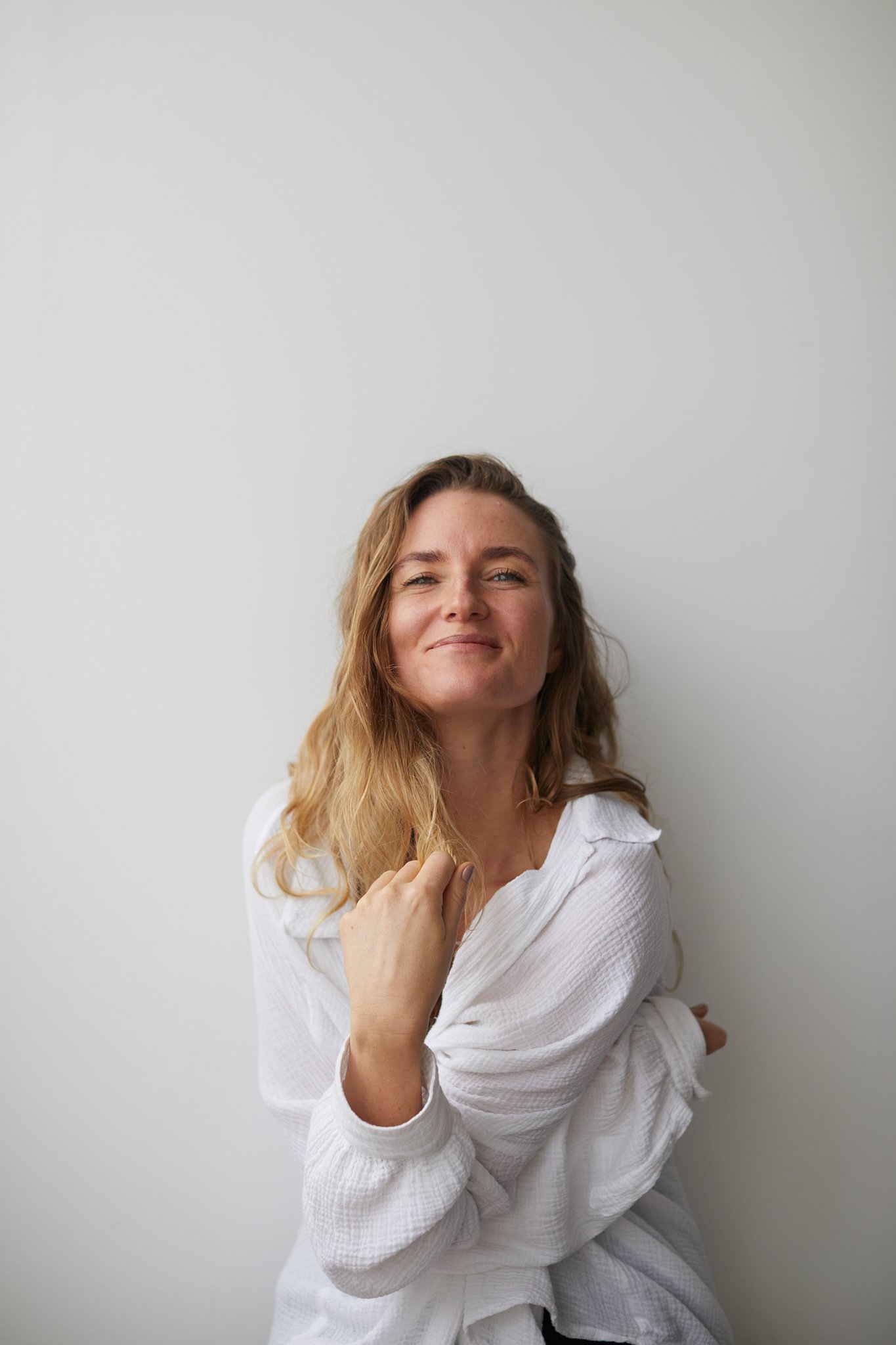 A woman with light brown hair, wearing a white shirt, smiling confidently, standing against a plain white background.