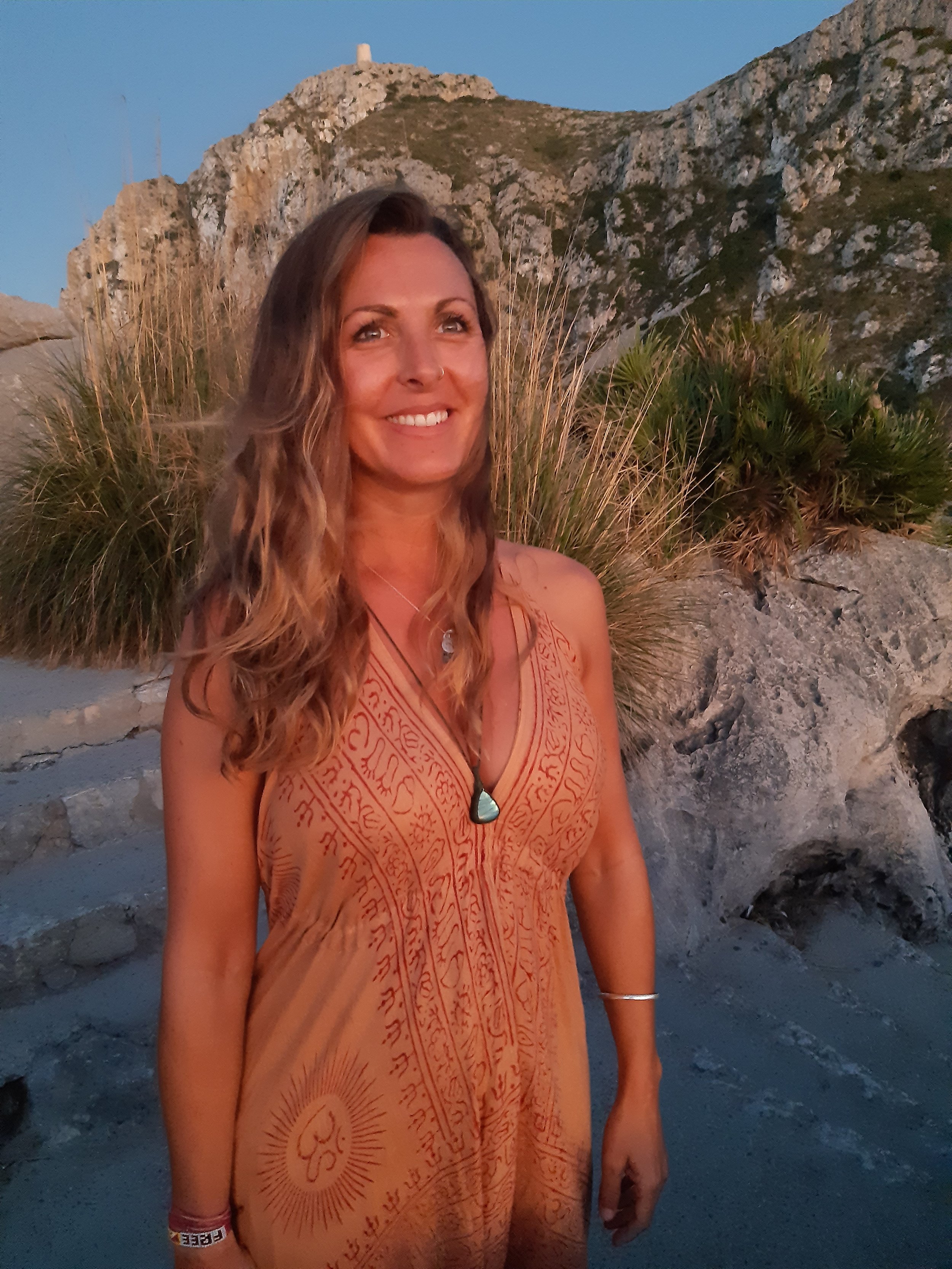A woman with long, wavy hair smiling outdoors on a rocky beach with desert plants and a mountain with a small white structure on top in the background.