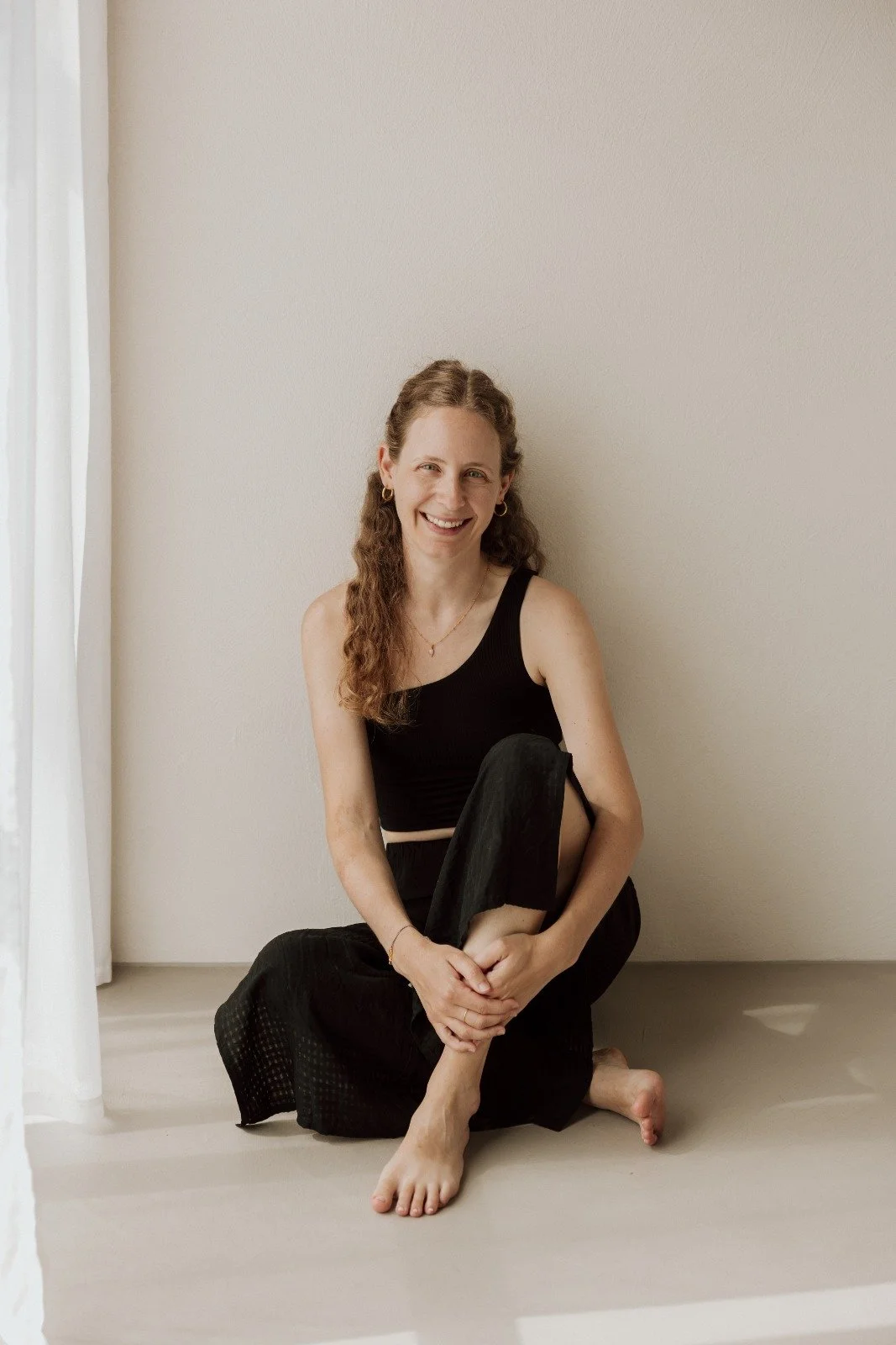 A woman with long, curly brown hair in black sleeveless top and pants, sitting on the floor and smiling, against a pale wall with a sheer curtain on the side.