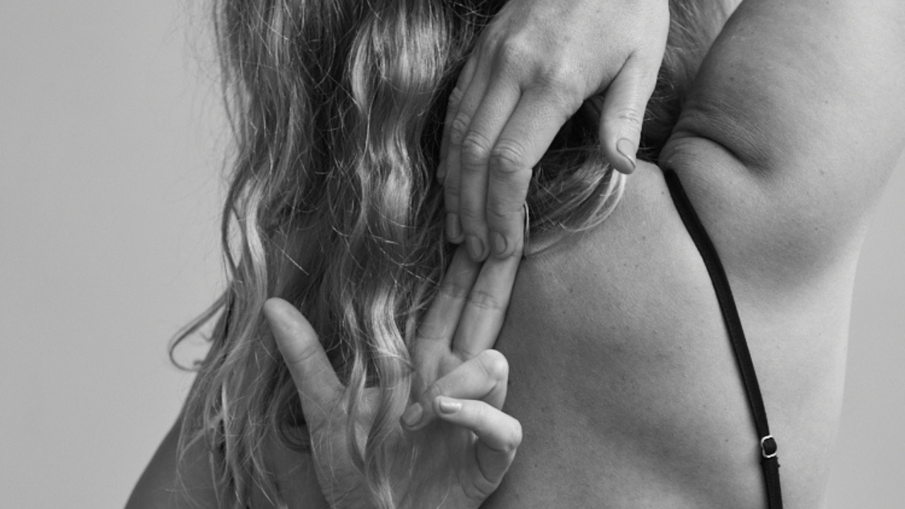 Close-up of a woman posing with her fingers in a peace sign near her face, showing her shoulder, with long wavy hair on a gray background.