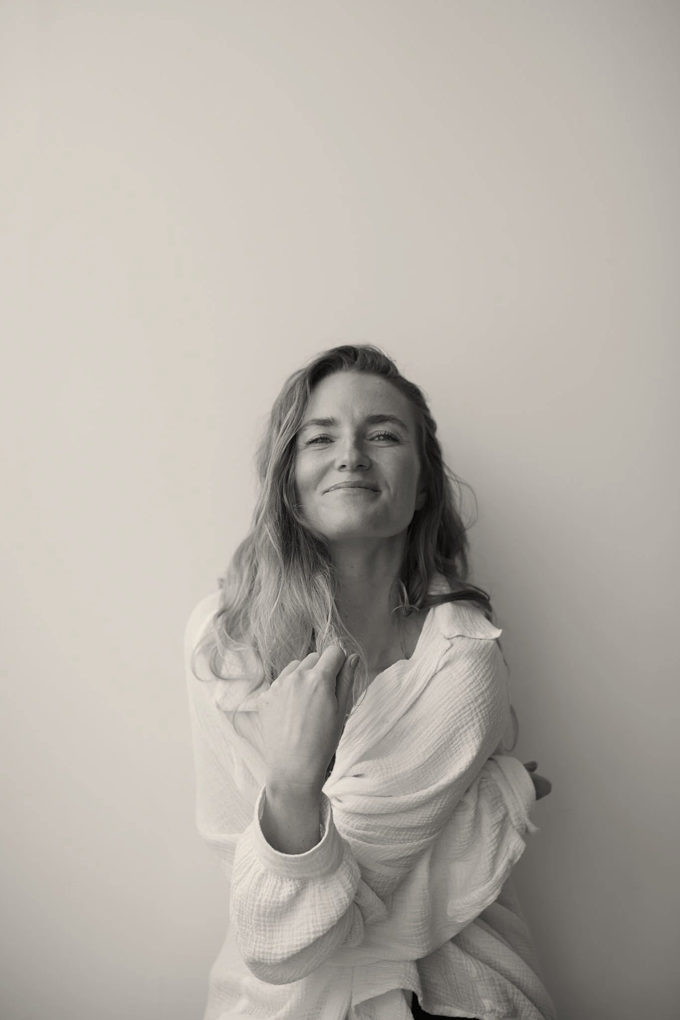 Black and white photo of a woman in a white robe, smiling gently, with one hand touching her chest and the other crossing her body, sitting against a plain wall.