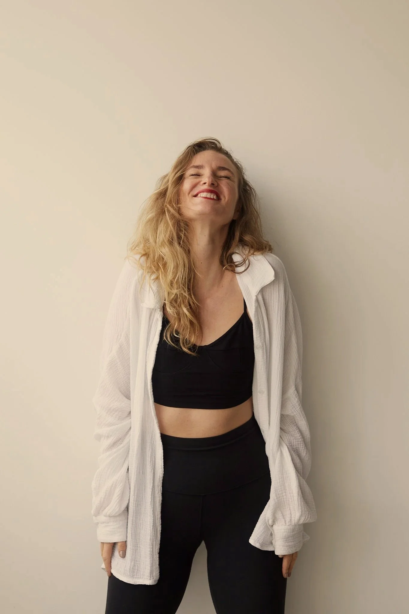 A woman standing against a plain light-colored wall, smiling with eyes closed, wearing a black crop top and black pants, with a white oversized cardigan.