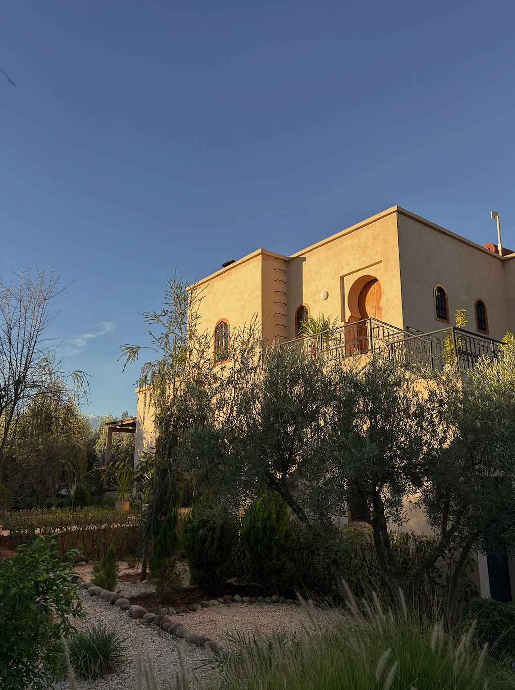 A modern two-story house with a beige exterior, arched windows, and a balcony, surrounded by landscaped greenery under a clear blue sky.