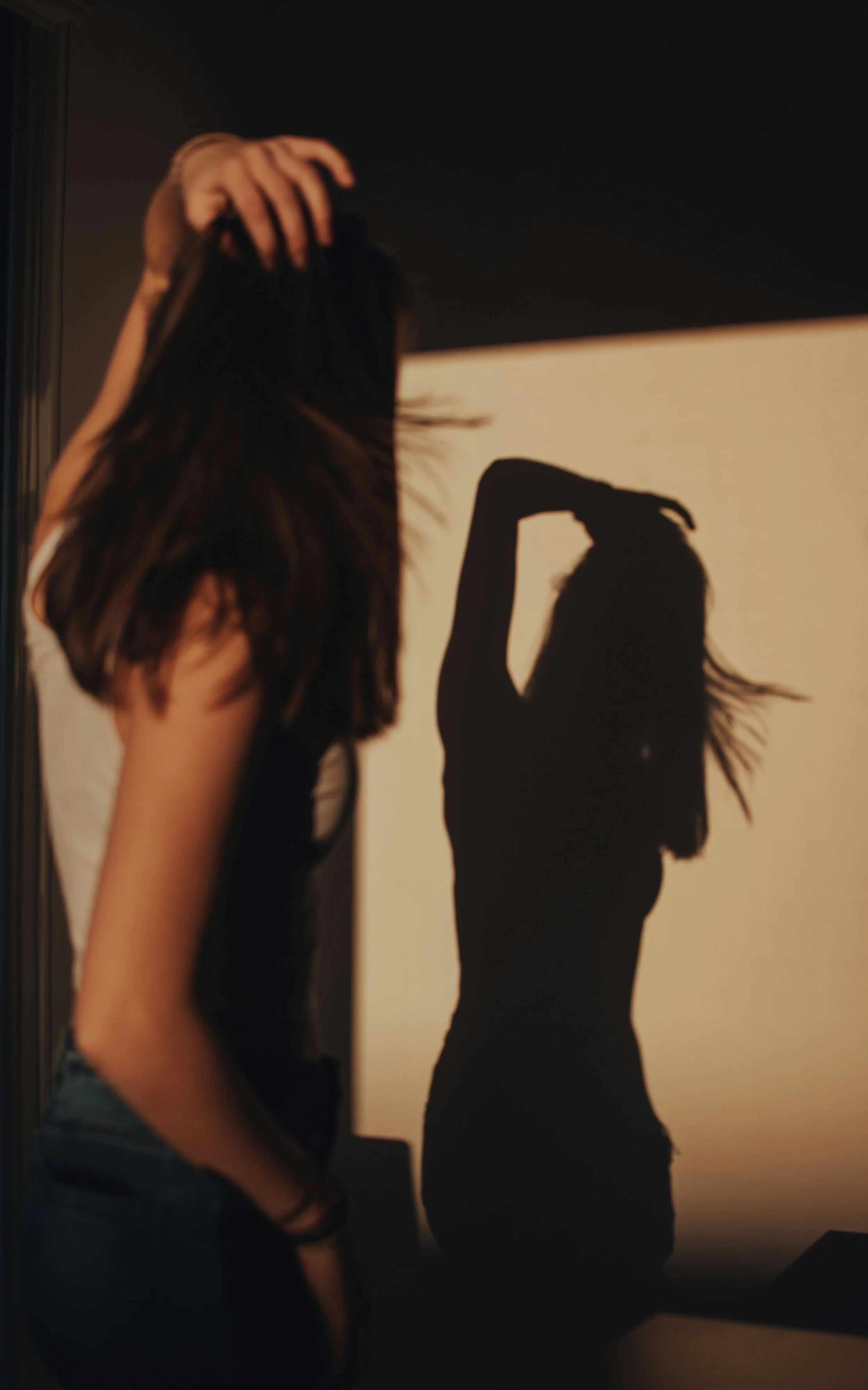 A woman with long hair takes a selfie in front of a wall with her shadow cast on it, showing a different pose with her hand on her head.