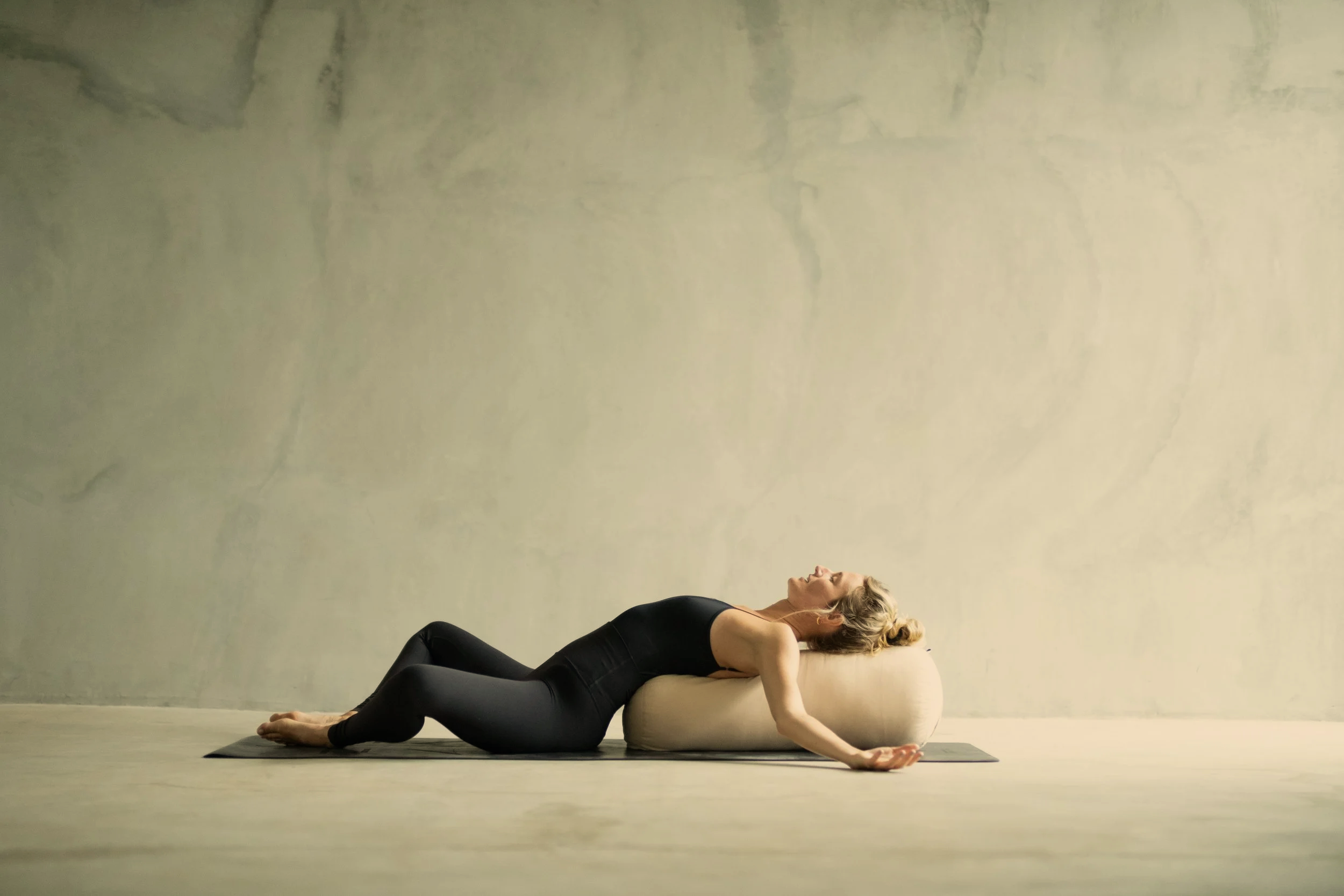 A woman practicing yoga on a mat against a plain wall, using a large cylindrical bolster for support, with her eyes closed in a relaxing pose.