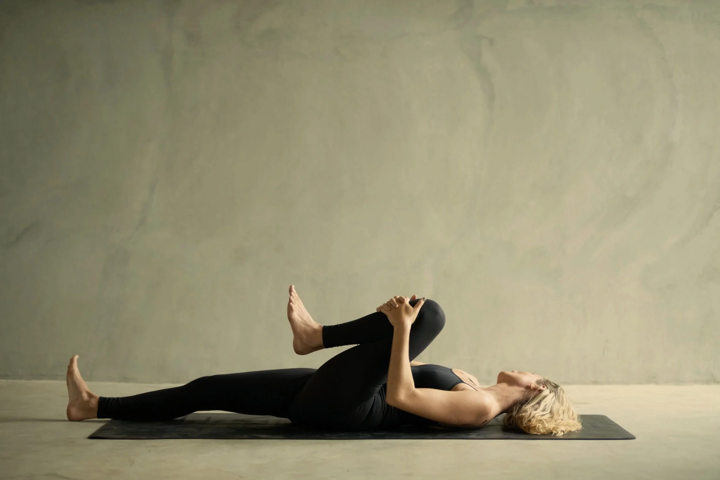 A woman practicing yoga on a mat against a plain wall, lying on her side with one arm supporting her head and the other hand holding her raised leg.