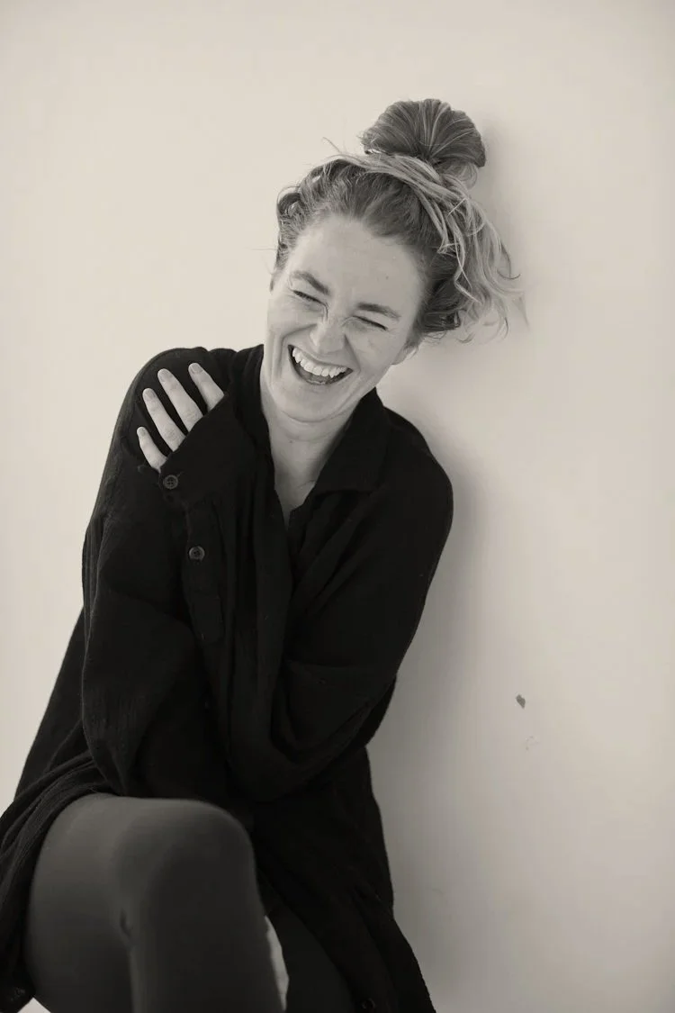 A woman with her hair in a messy bun, wearing a dark jacket, laughing while leaning against a plain wall.