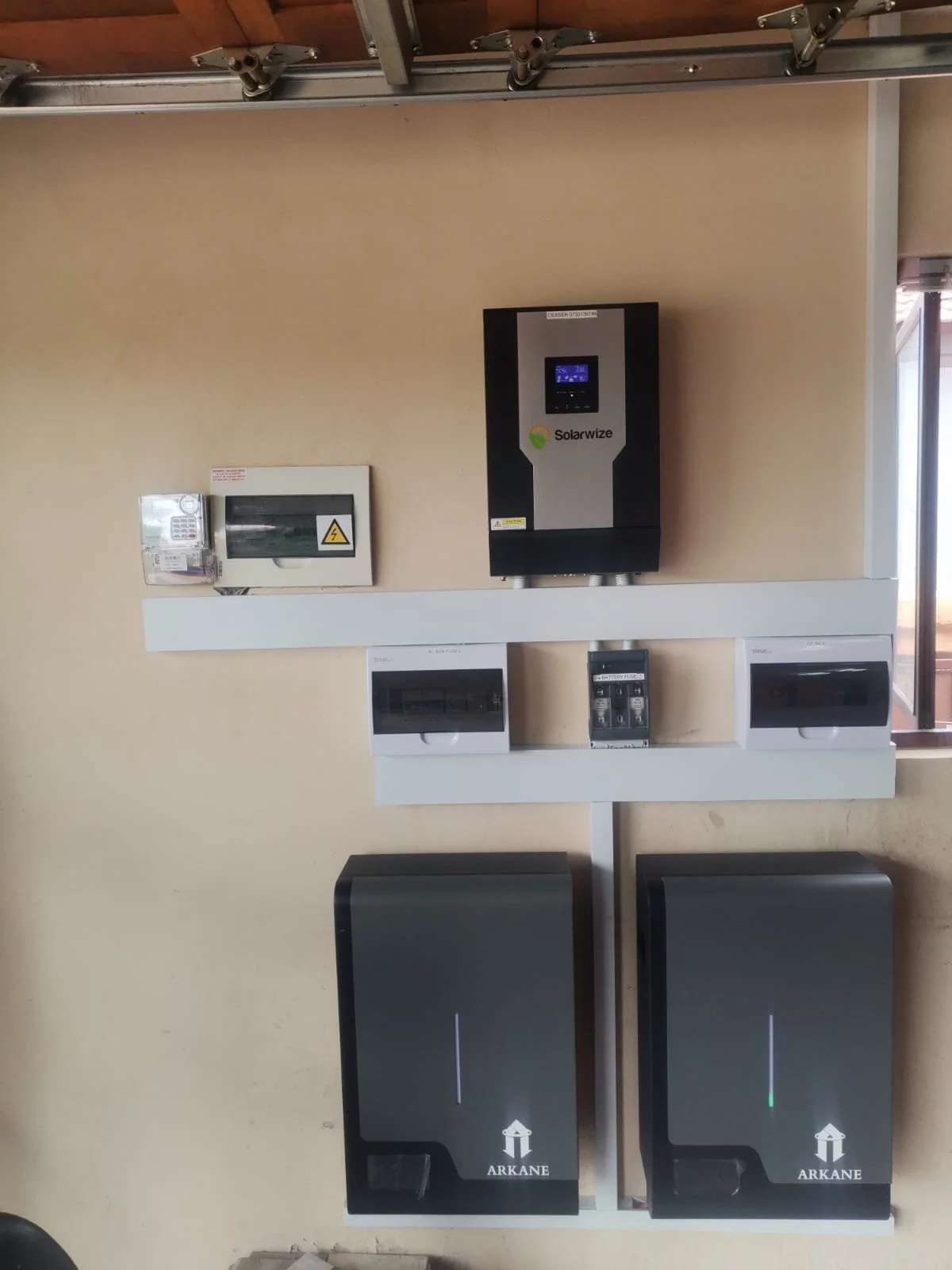 Wall-mounted solar energy equipment and electrical panels, including solar inverter, circuit breaker, battery storage, and solar energy storage boxes labeled 'ARKANE' in a room with a window to the right.